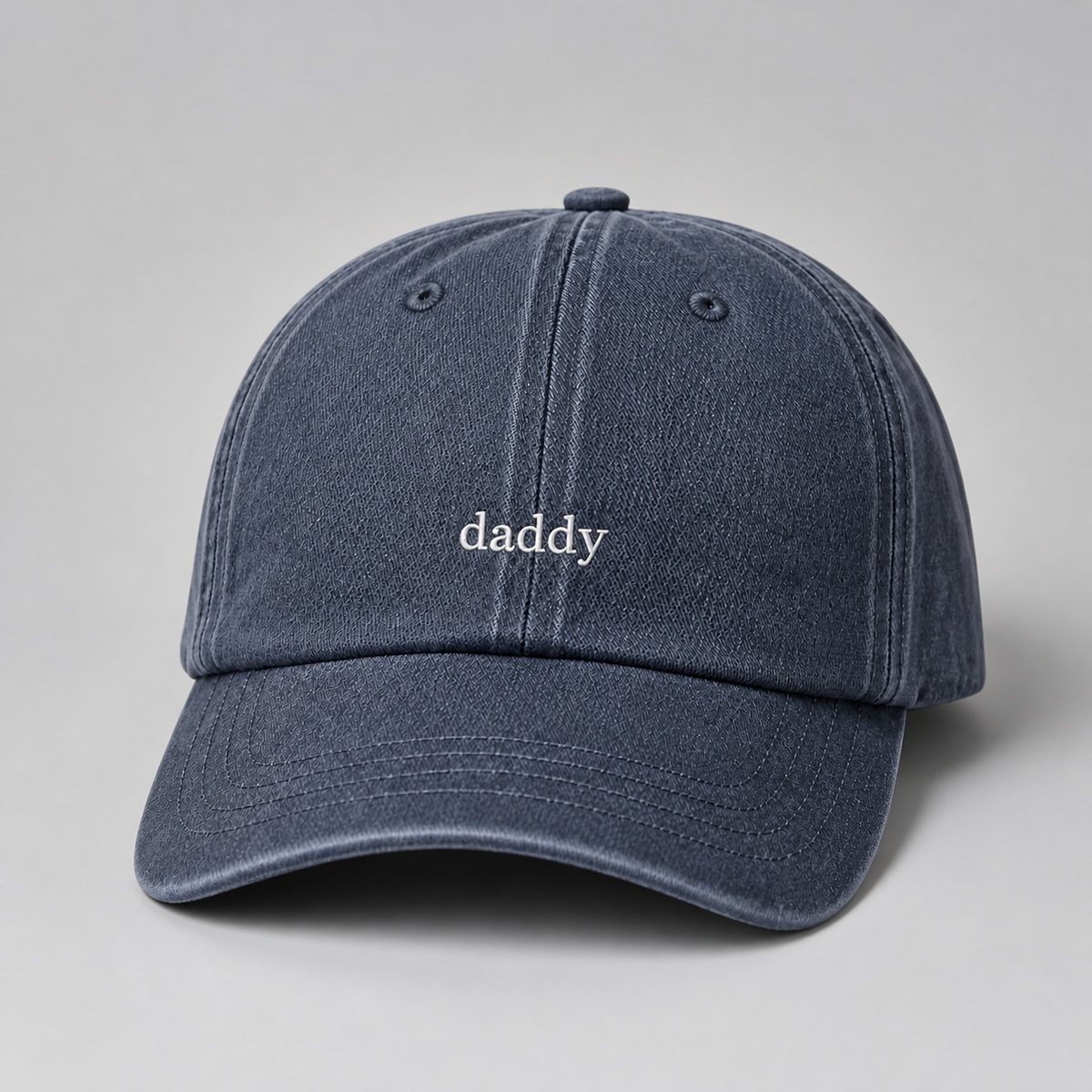 A washed navy blue denim baseball cap with the word 'daddy' embroidered in white lowercase letters on the front, set against a neutral gray background.