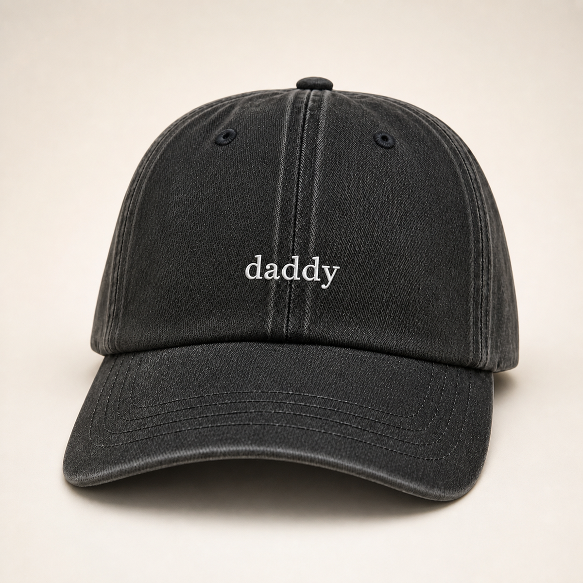 A dark washed black baseball cap with the word 'daddy' embroidered in white lowercase letters on the front, set against a plain light beige background.