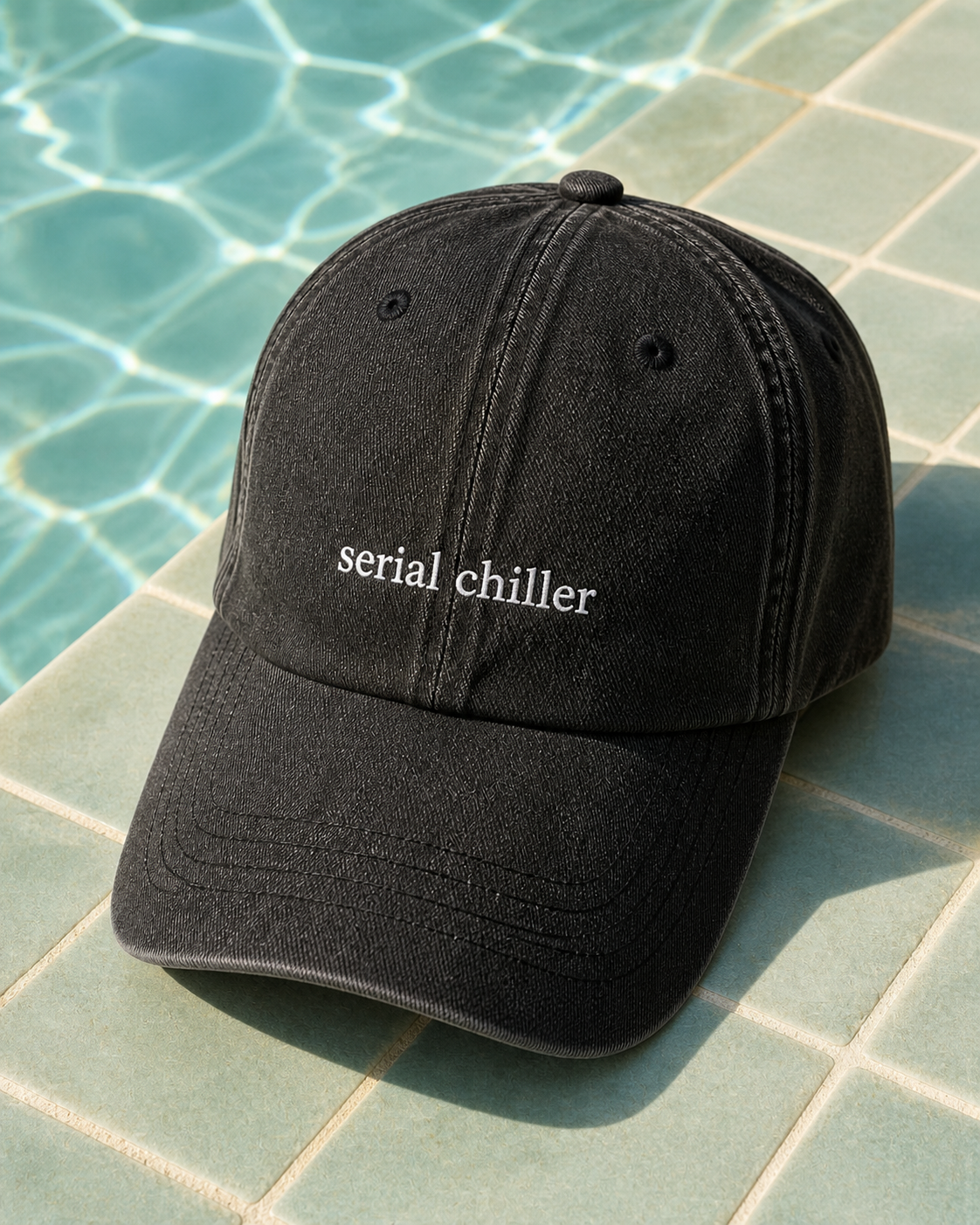 A black baseball cap with the words 'serial chiller' embroidered in white on the front, resting on a tiled surface next to a swimming pool.