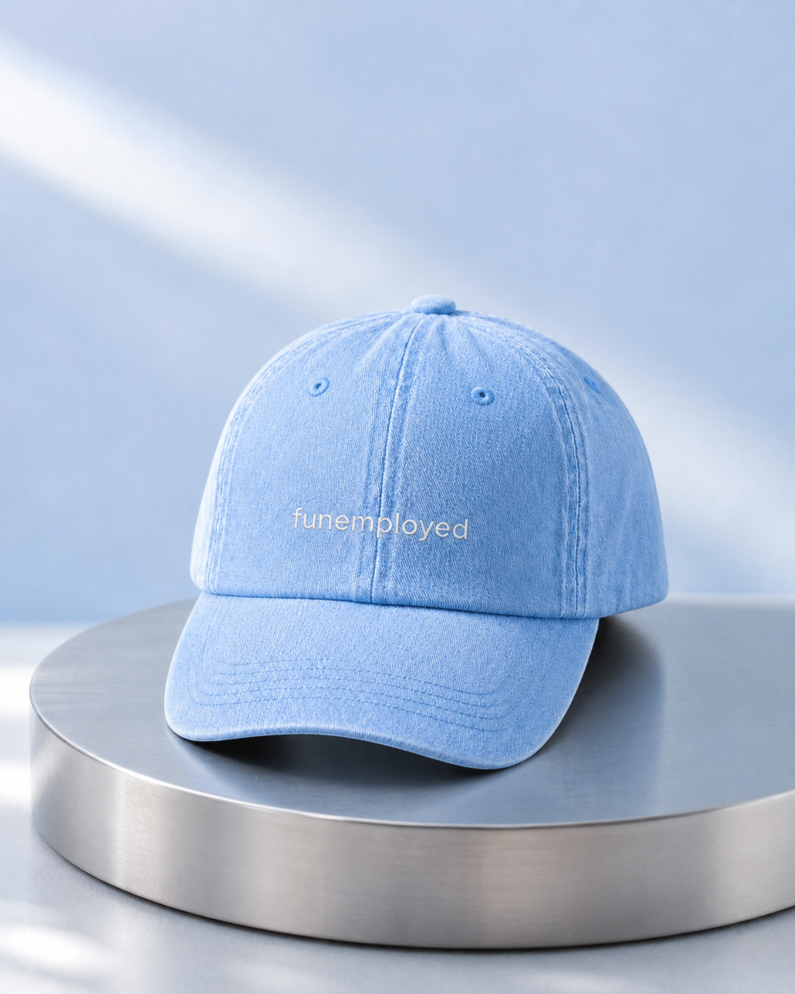 A light blue denim baseball cap with the word 'funemployed' embroidered in white on the front, displayed on a round metallic pedestal against a soft light blue background.