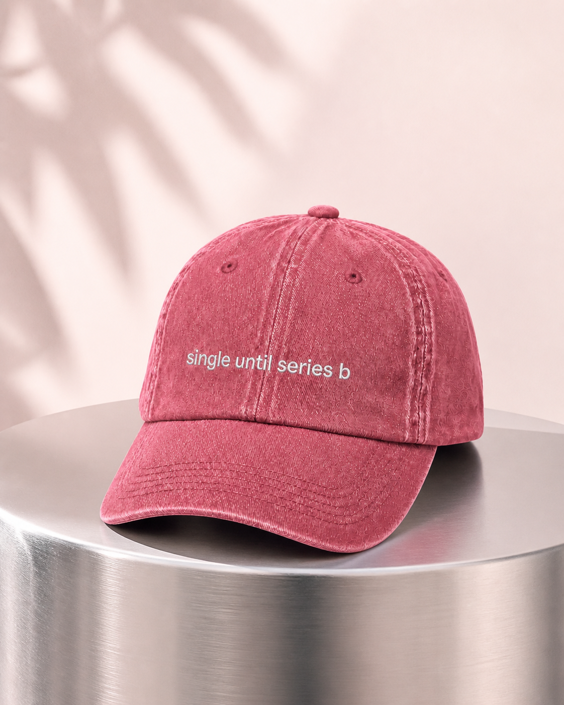 A dusty rose pink washed cotton baseball cap with white embroidered text reading 'single until series b' on the front, resting on a metallic silver cylindrical pedestal against a warm beige background with soft palm leaf shadows.