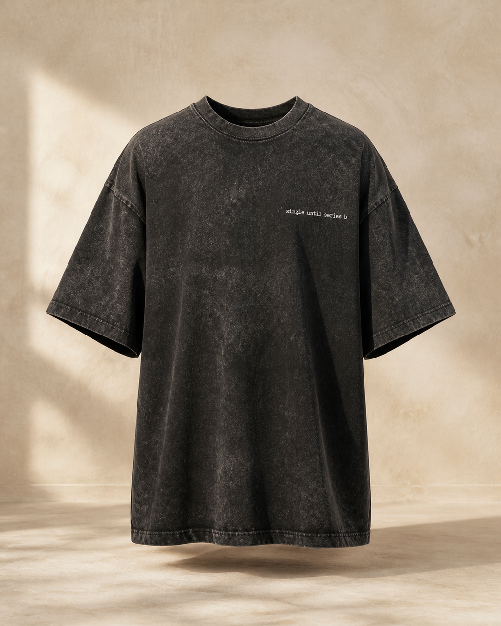 A washed black oversized short-sleeve t-shirt with a small white text print on the left chest, displayed against a textured beige wall with soft window shadows.