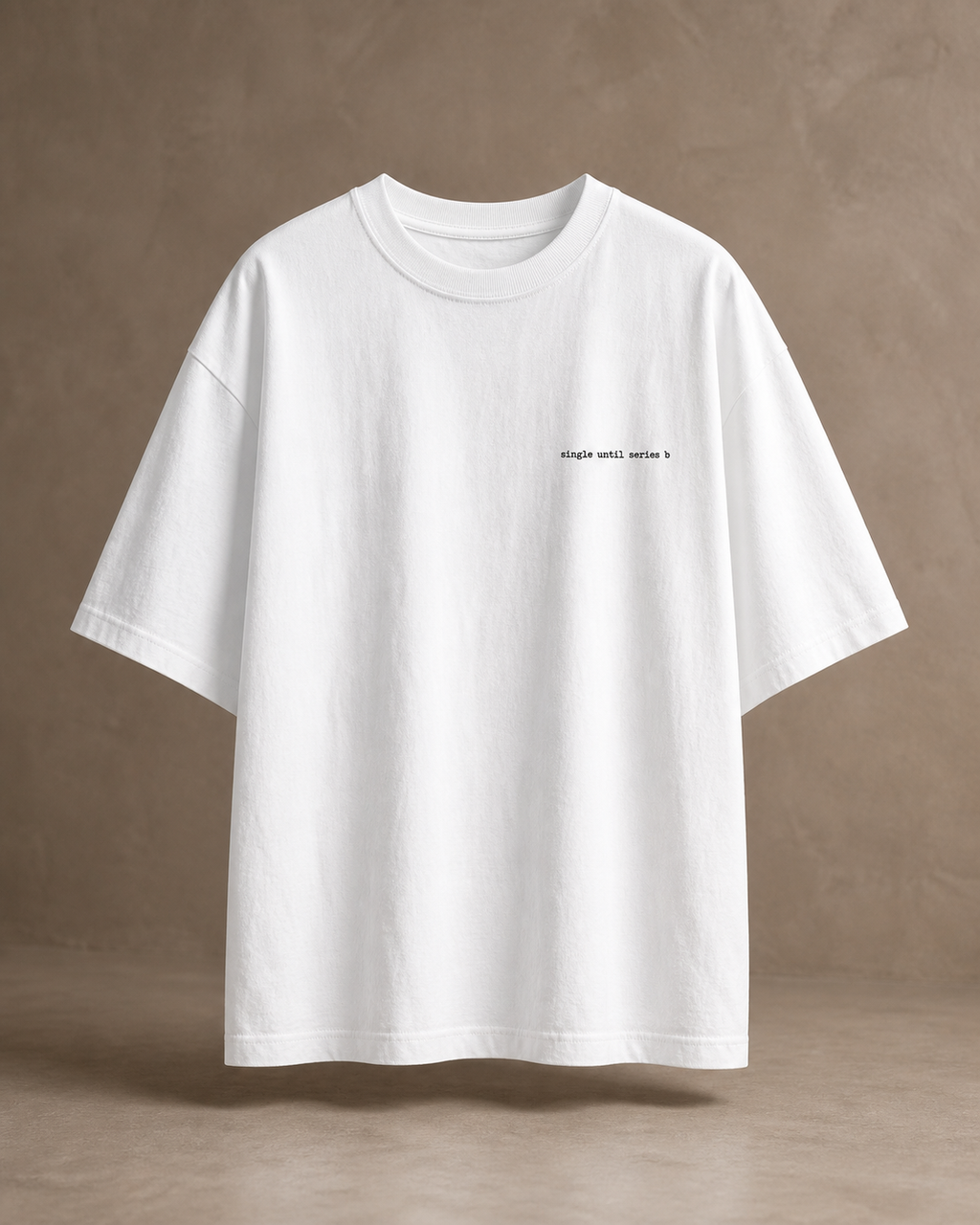 An oversized white short-sleeve t-shirt with a small black text print on the left chest, displayed against a textured beige wall with soft window shadows.