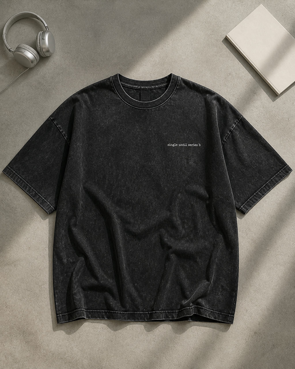 A black washed short-sleeve t-shirt with small white text on the left chest, laid flat on a textured gray surface with headphones and a book as props.