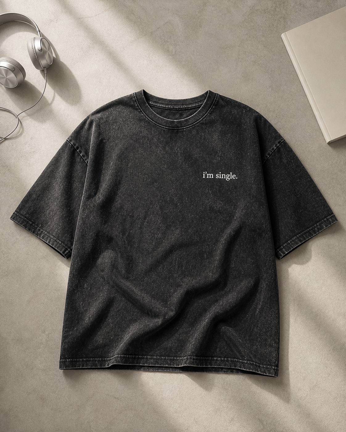 A washed black short-sleeve t-shirt with 'i'm single.' printed in white on the left chest, laid flat on a textured gray surface with styled props including headphones and a book.