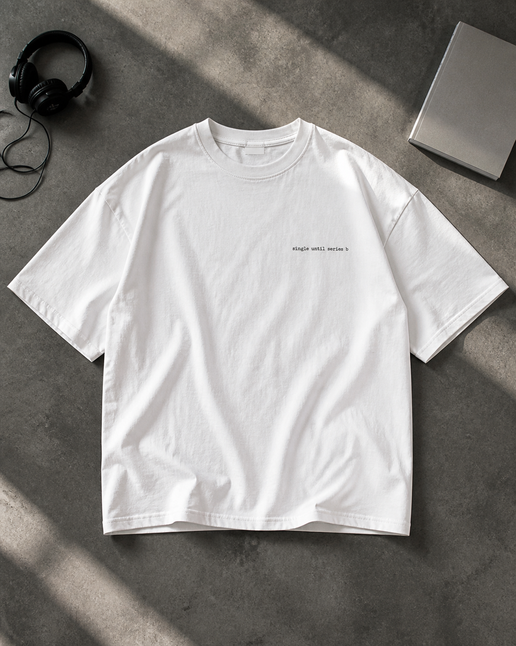 A white short-sleeve t-shirt with small black text on the left chest, laid flat on a textured grey surface with headphones and a book as props.