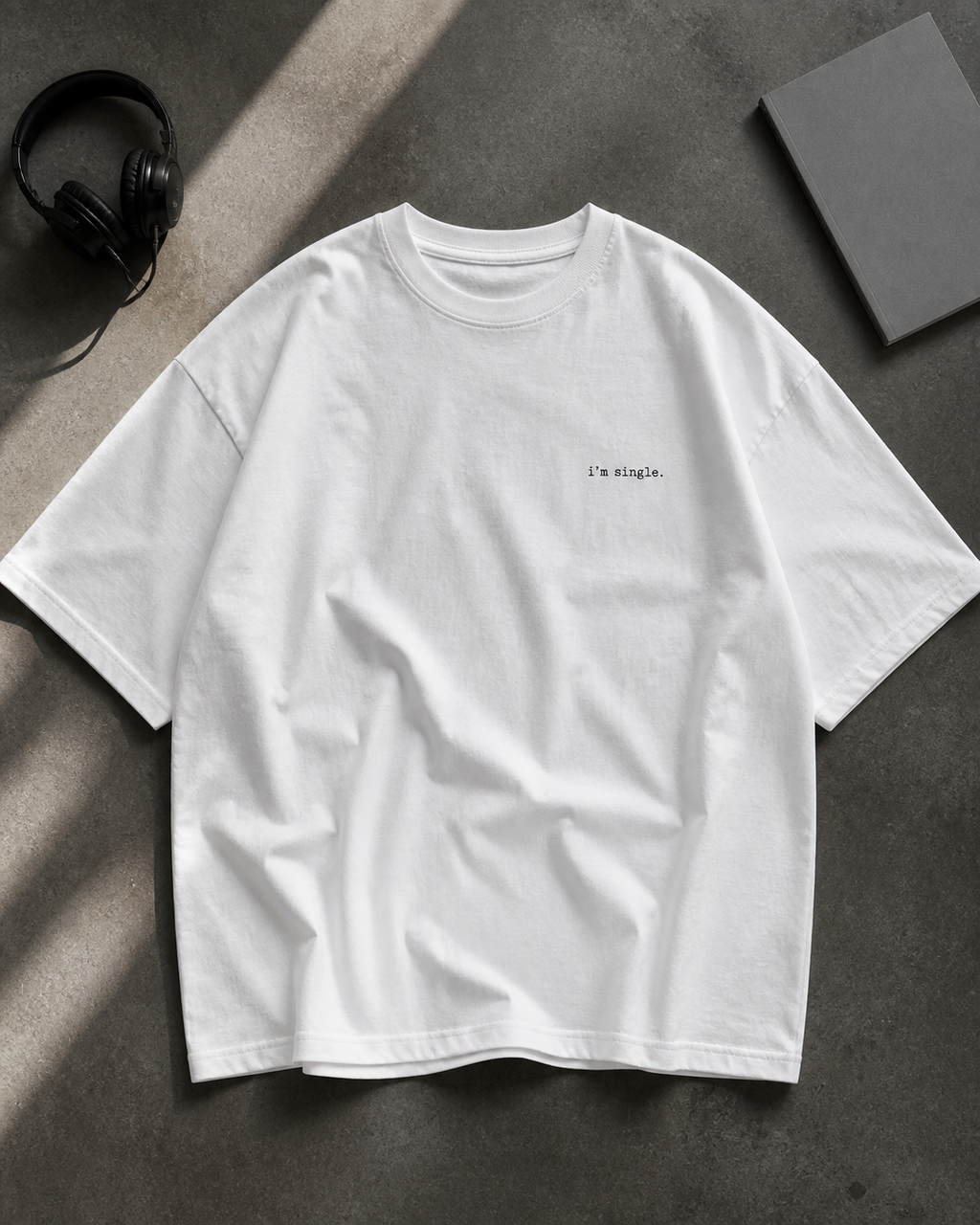 A white short-sleeve t-shirt with 'i'm single.' printed in black on the left chest, laid flat on a textured grey surface with headphones and a book.