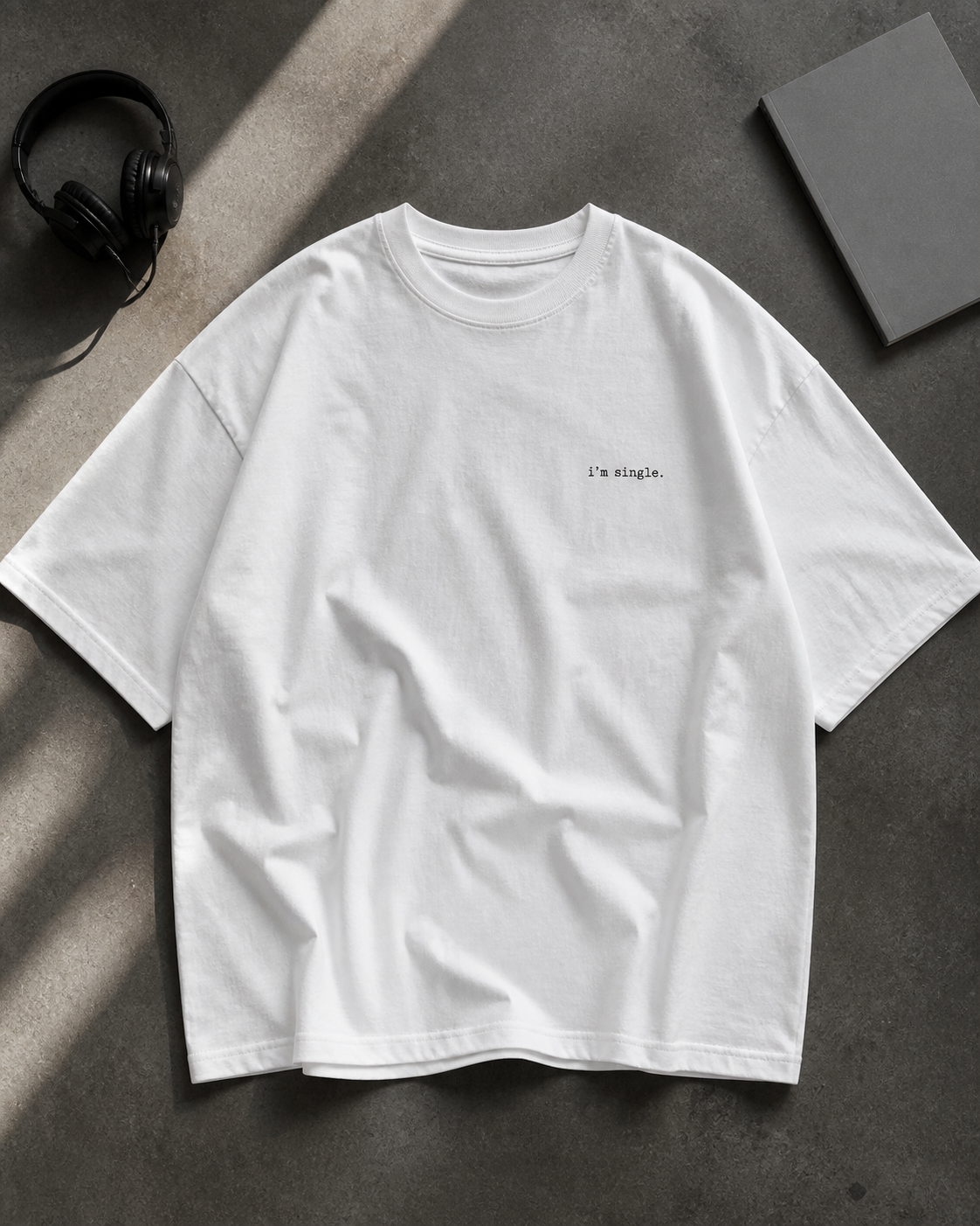 A white short-sleeve t-shirt with 'i'm single.' printed in black on the left chest, laid flat on a textured grey surface with headphones and a book.