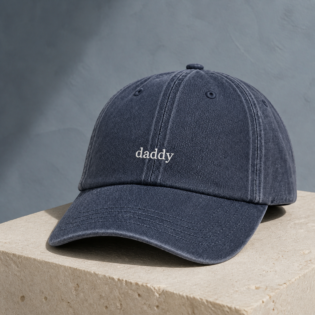 A dark washed denim baseball cap with the word 'daddy' embroidered in white on the front, resting on a light beige stone block against a textured gray background.