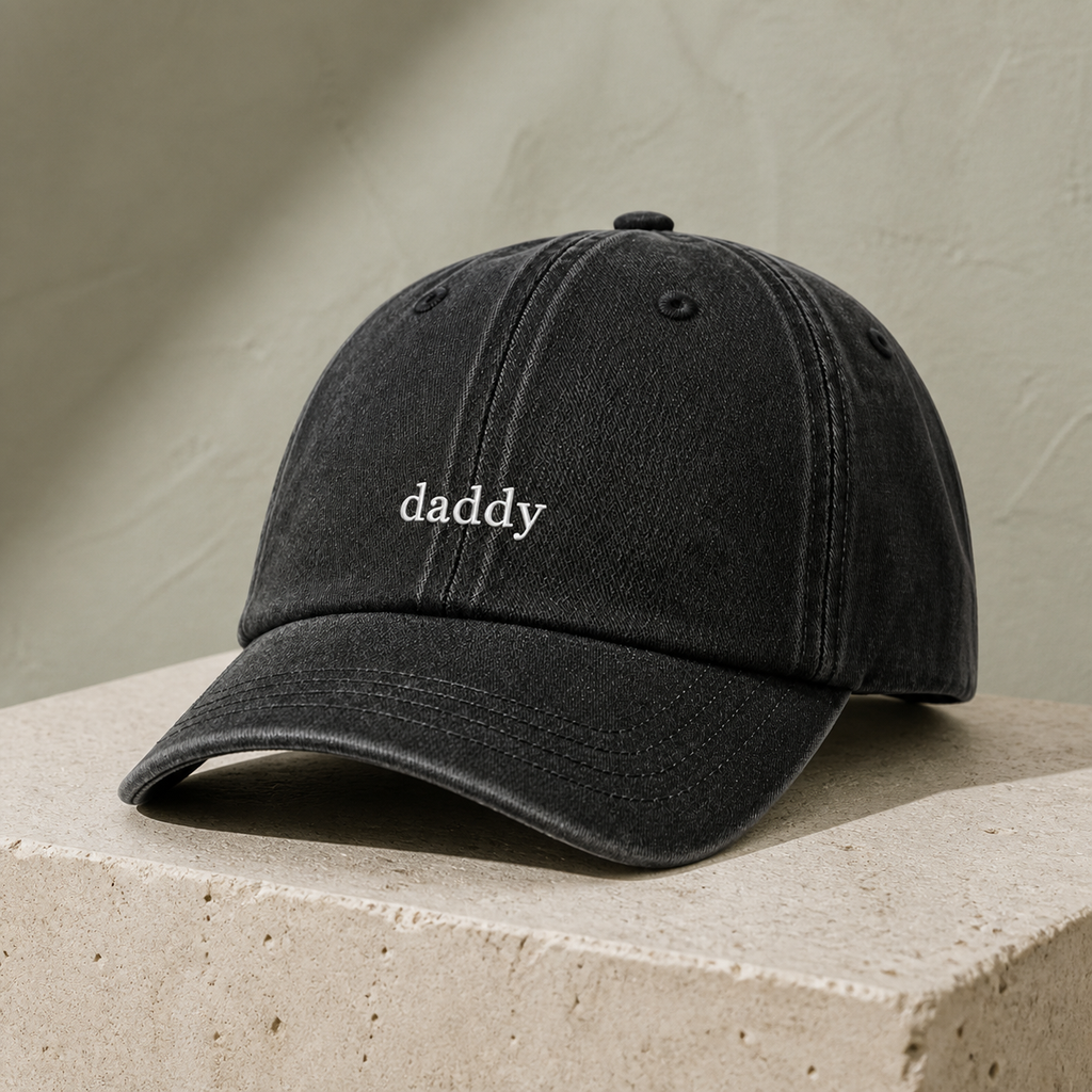 A black washed baseball cap with the word 'daddy' embroidered in white on the front, resting on a textured beige stone block against a neutral beige wall.