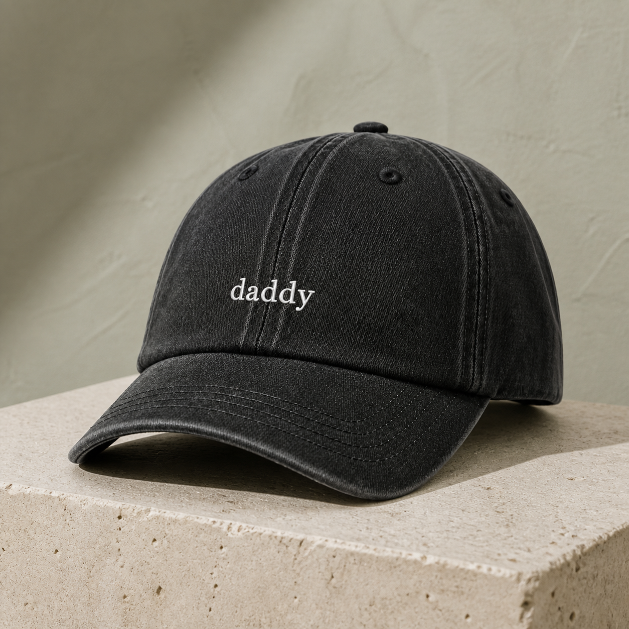 A black washed baseball cap with the word 'daddy' embroidered in white on the front, resting on a textured beige stone block against a neutral beige wall.
