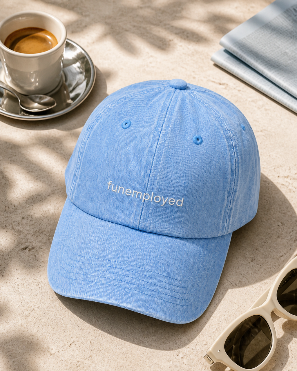 A light blue cotton baseball cap with the word 'funemployed' embroidered in white on the front, styled on a beige stone surface with a cup of coffee, sunglasses, and a folded blue towel nearby.