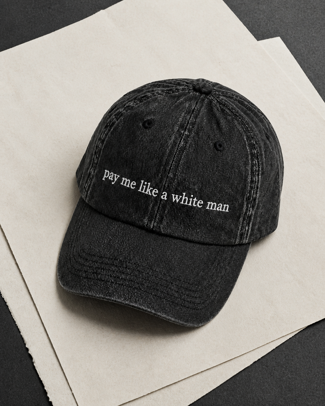 A washed black baseball cap with white embroidered text reading 'pay me like a white man' resting on stacked off-white paper sheets over a dark surface.