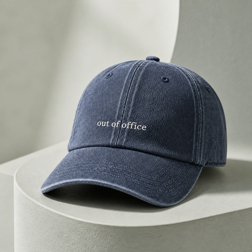 A navy blue washed cotton baseball cap with 'out of office' embroidered in white on the front, resting on a light gray curved pedestal.