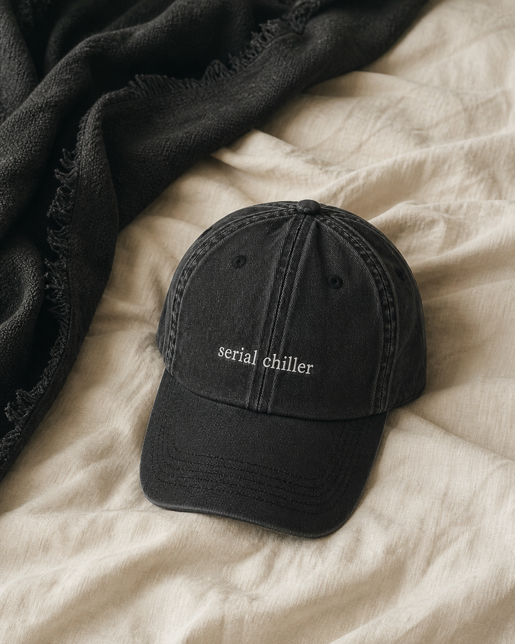A black washed baseball cap with 'serial chiller' embroidered in white on the front, styled on a beige linen surface next to a dark charcoal throw blanket.