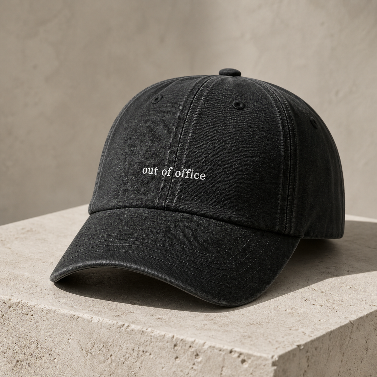 A washed black baseball cap with 'out of office' embroidered in white on the front, resting on a textured beige stone pedestal against a matching beige background.