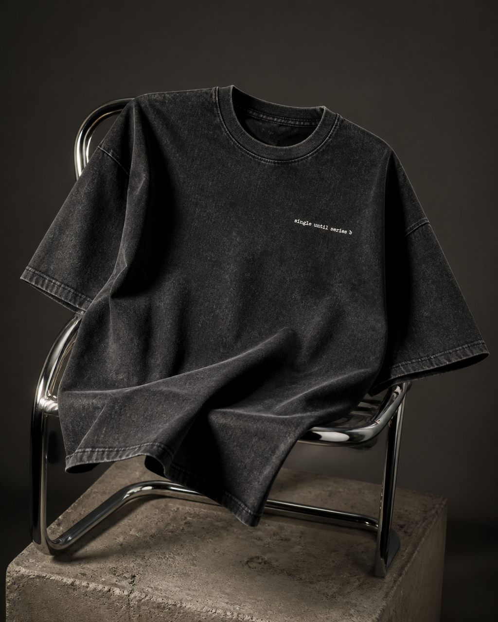 A washed black short-sleeve t-shirt with small white text on the left chest, draped over a modern metal chair on a concrete block against a dark studio background.
