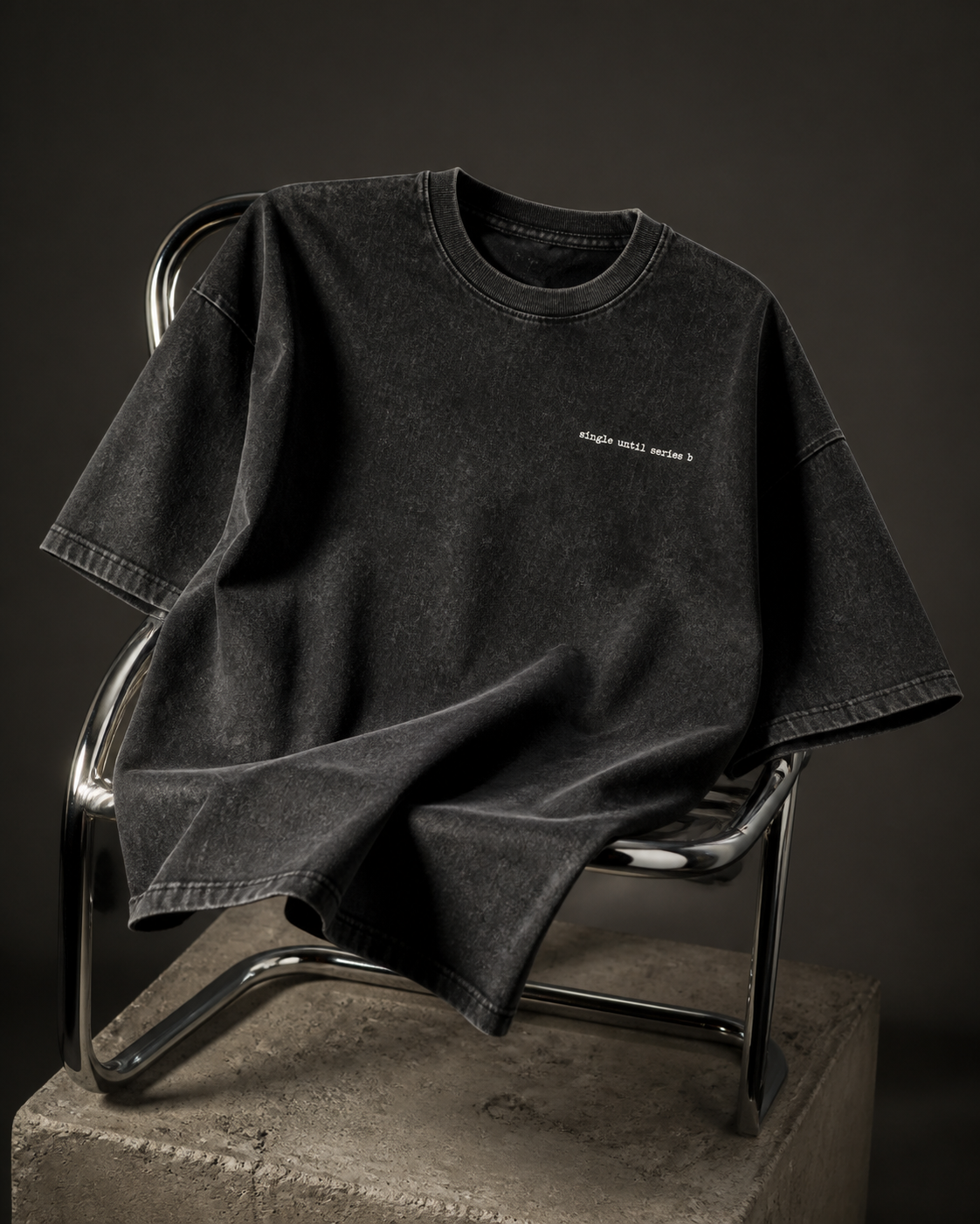 A washed black short-sleeve t-shirt with small white text on the left chest, draped over a modern metal chair on a concrete block against a dark studio background.