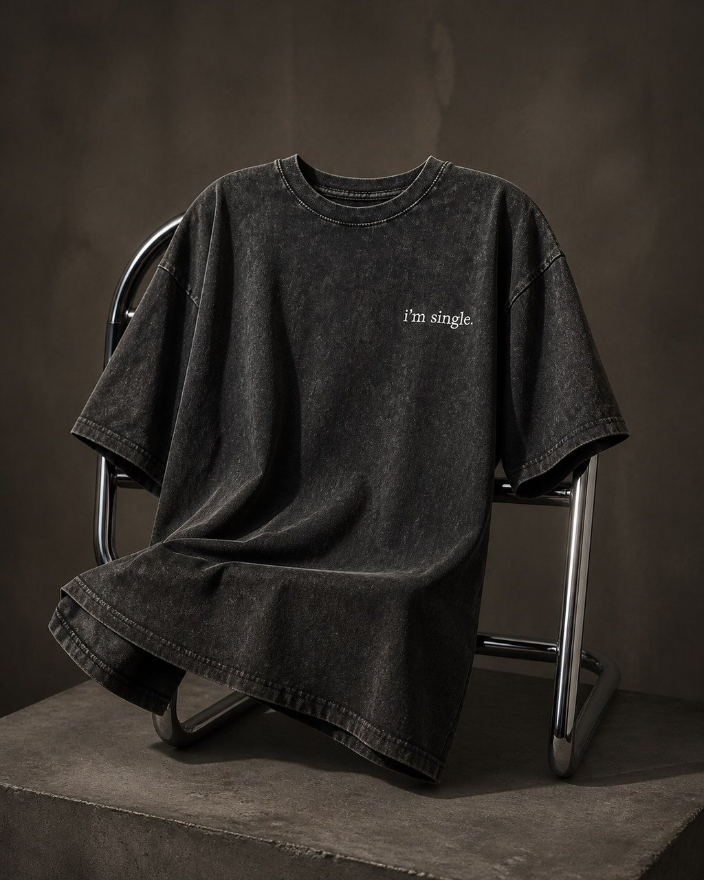 A washed black oversized short-sleeve t-shirt with the text 'I'm single.' printed in white on the left chest, draped over a metal chair on a concrete platform against a dark textured background.
