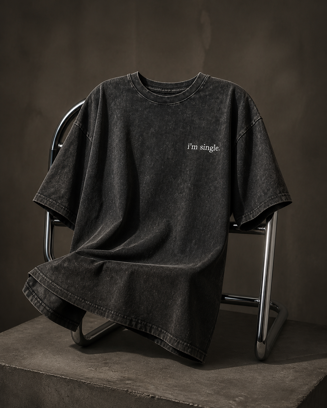 A washed black oversized short-sleeve t-shirt with the text 'I'm single.' printed in white on the left chest, draped over a metal chair on a concrete platform against a dark textured background.
