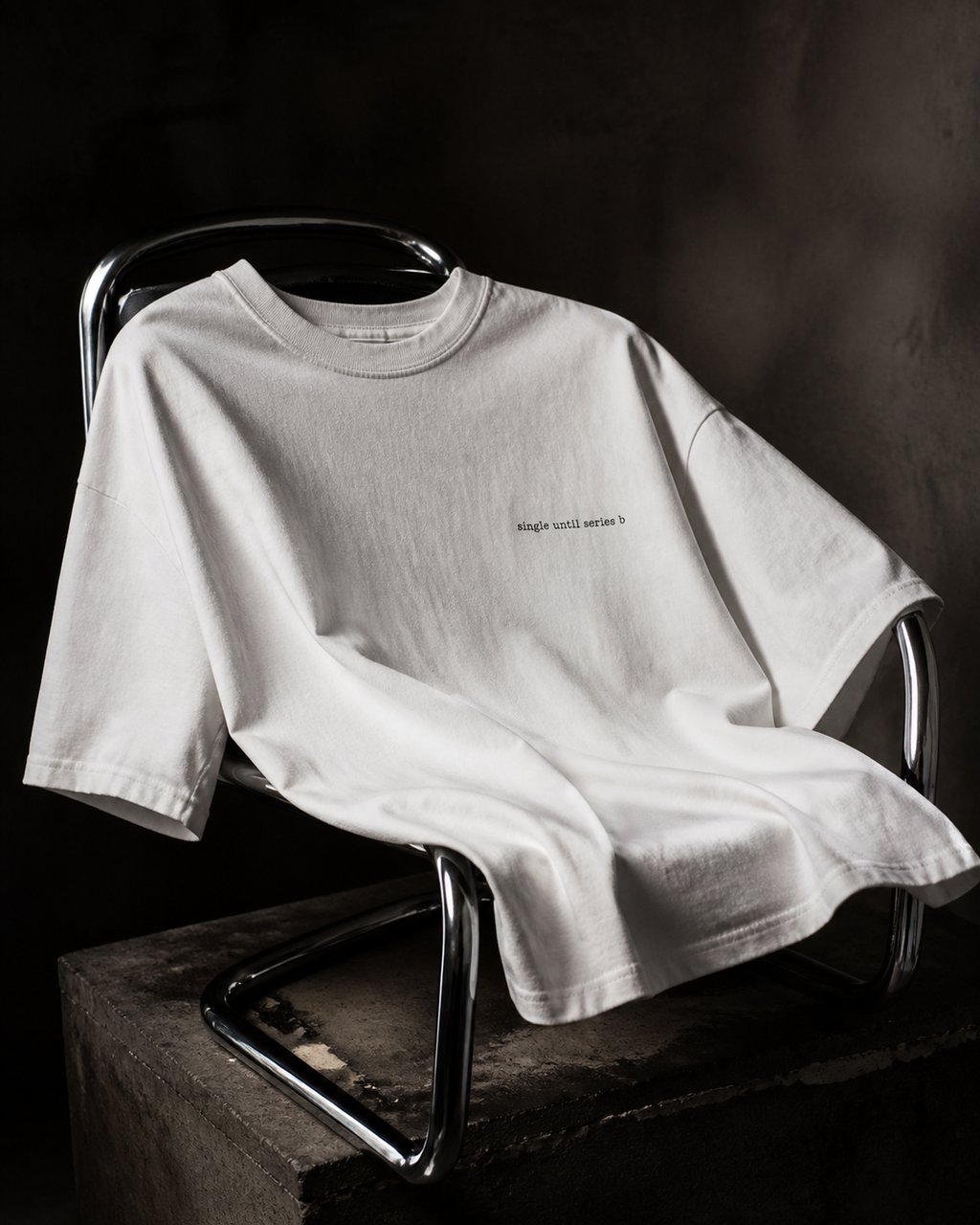 A white short-sleeve t-shirt with small black text on the left chest, draped over a modern metal chair on a concrete block against a dark studio background.