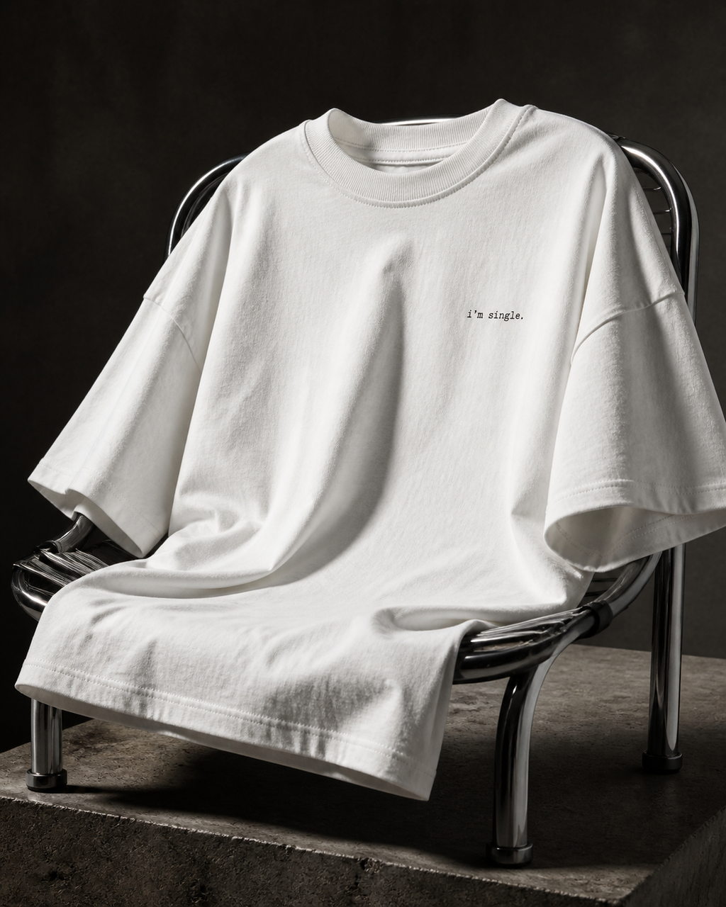 A white oversized short-sleeve t-shirt with 'i'm single.' printed in black on the left chest, draped over a metal chair on a concrete platform against a dark textured background.