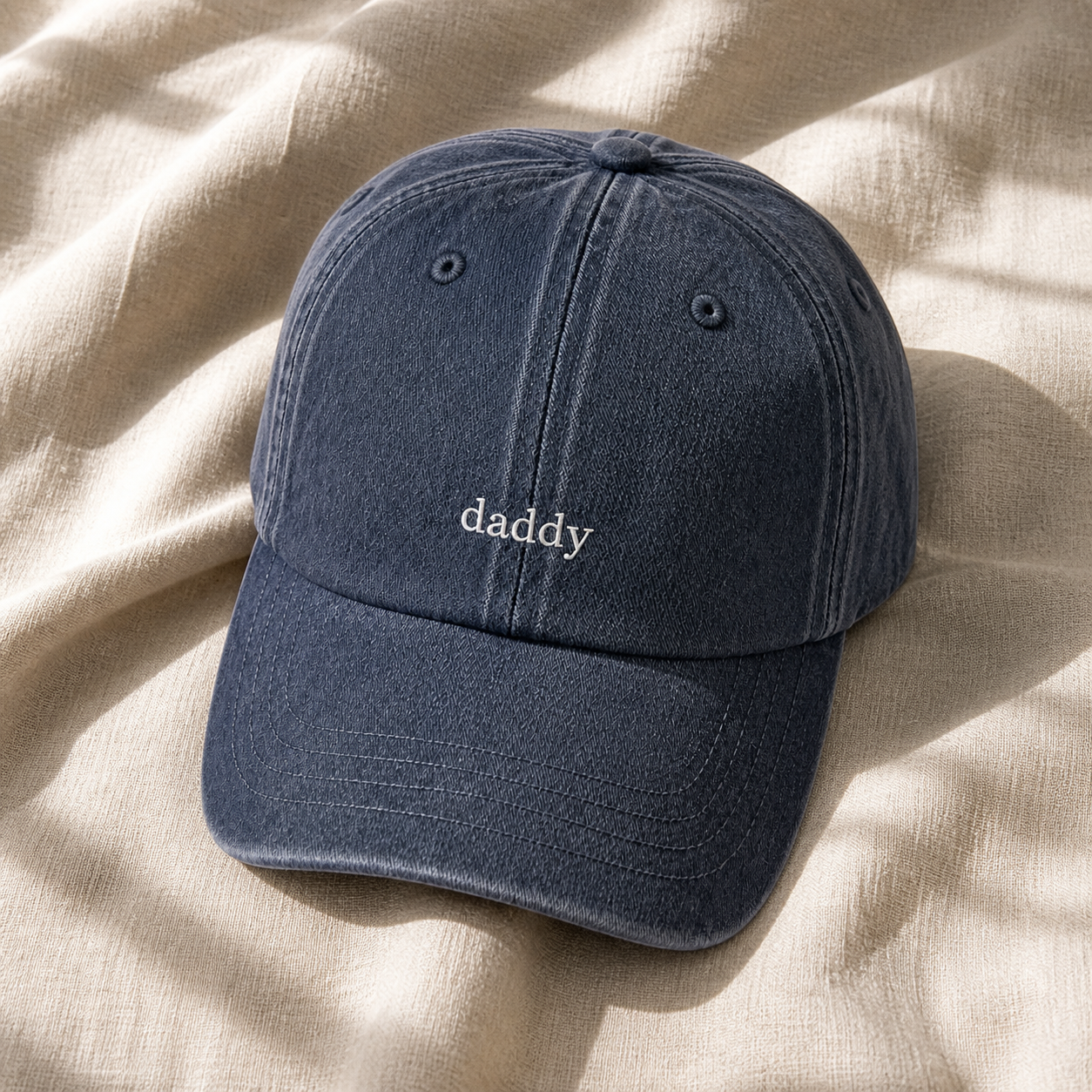 A dark navy blue washed cotton baseball cap with the word 'daddy' embroidered in white lowercase letters on the front, resting on a beige linen fabric background with soft leaf shadows.