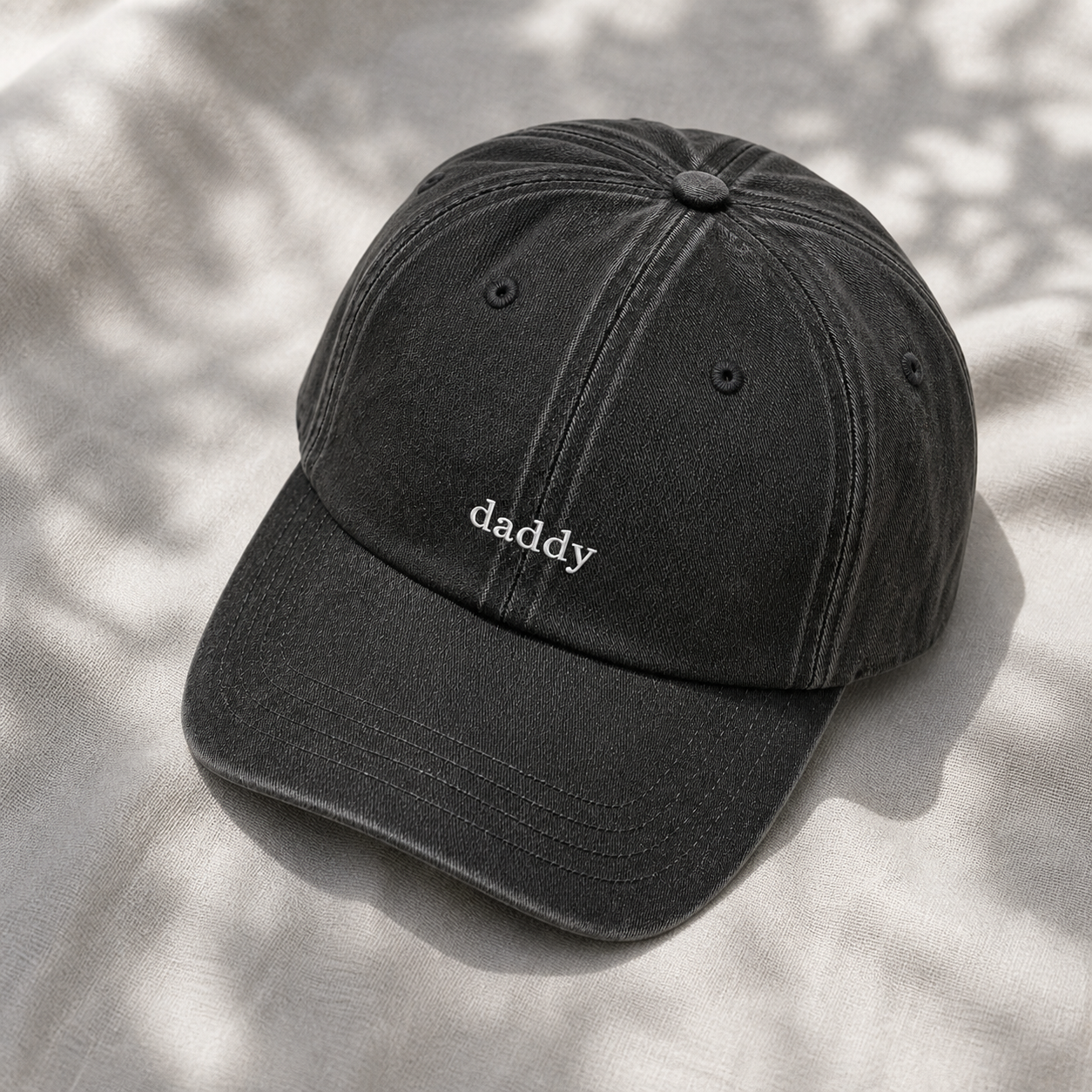 A washed black baseball cap with the word 'daddy' embroidered in white on the front, resting on a light gray textured fabric with soft leaf shadows.