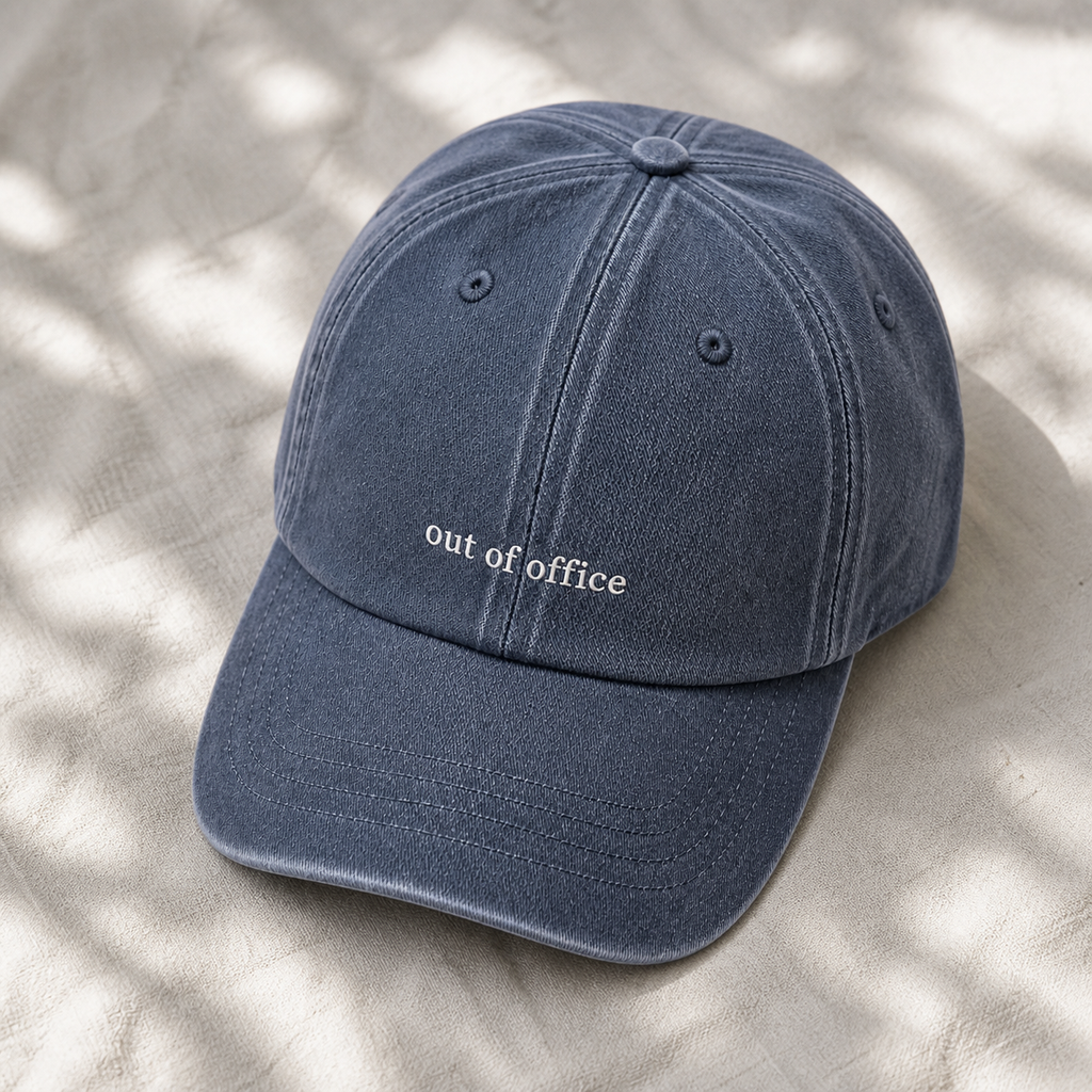 A washed navy blue cotton baseball cap with 'out of office' embroidered in white on the front, resting on a textured beige fabric with soft leaf shadows.