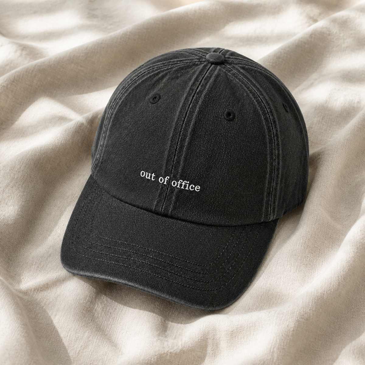 A washed black cotton baseball cap with 'out of office' embroidered in white on the front, resting on a draped beige fabric background.
