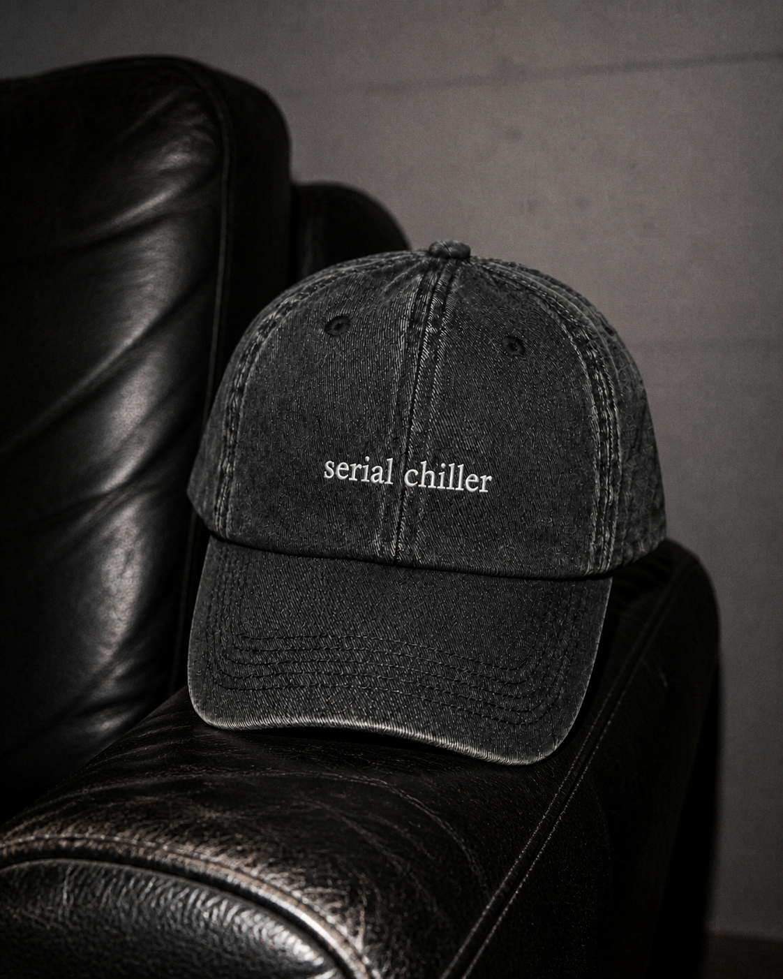 A washed black baseball cap with the words 'serial chiller' embroidered in white on the front, resting on a black leather armchair against a dark gray wall.