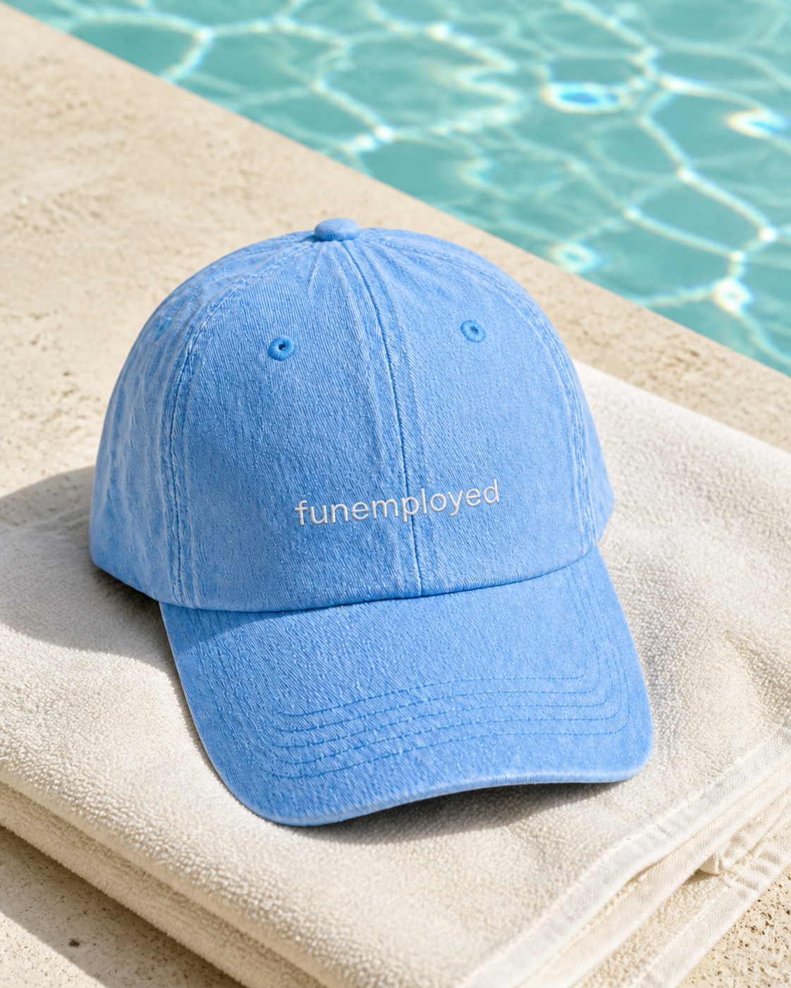 A light blue baseball cap with the word 'funemployed' embroidered on the front, resting on a beige towel beside a swimming pool.
