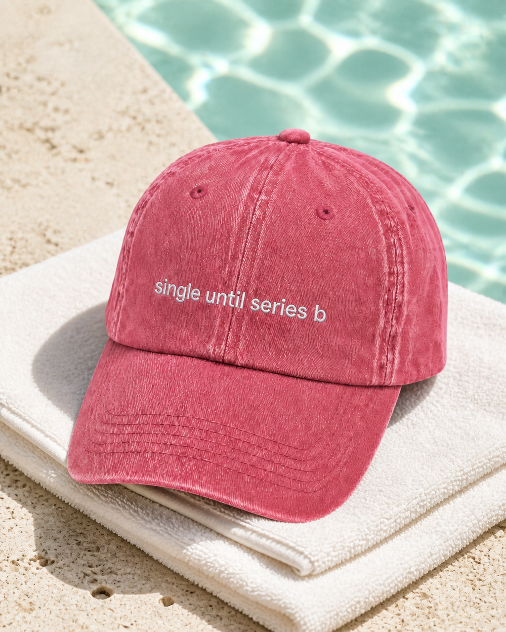 A dusty rose pink cotton cap with the phrase 'single until series b' embroidered in white on the front, resting on a white towel next to a swimming pool.