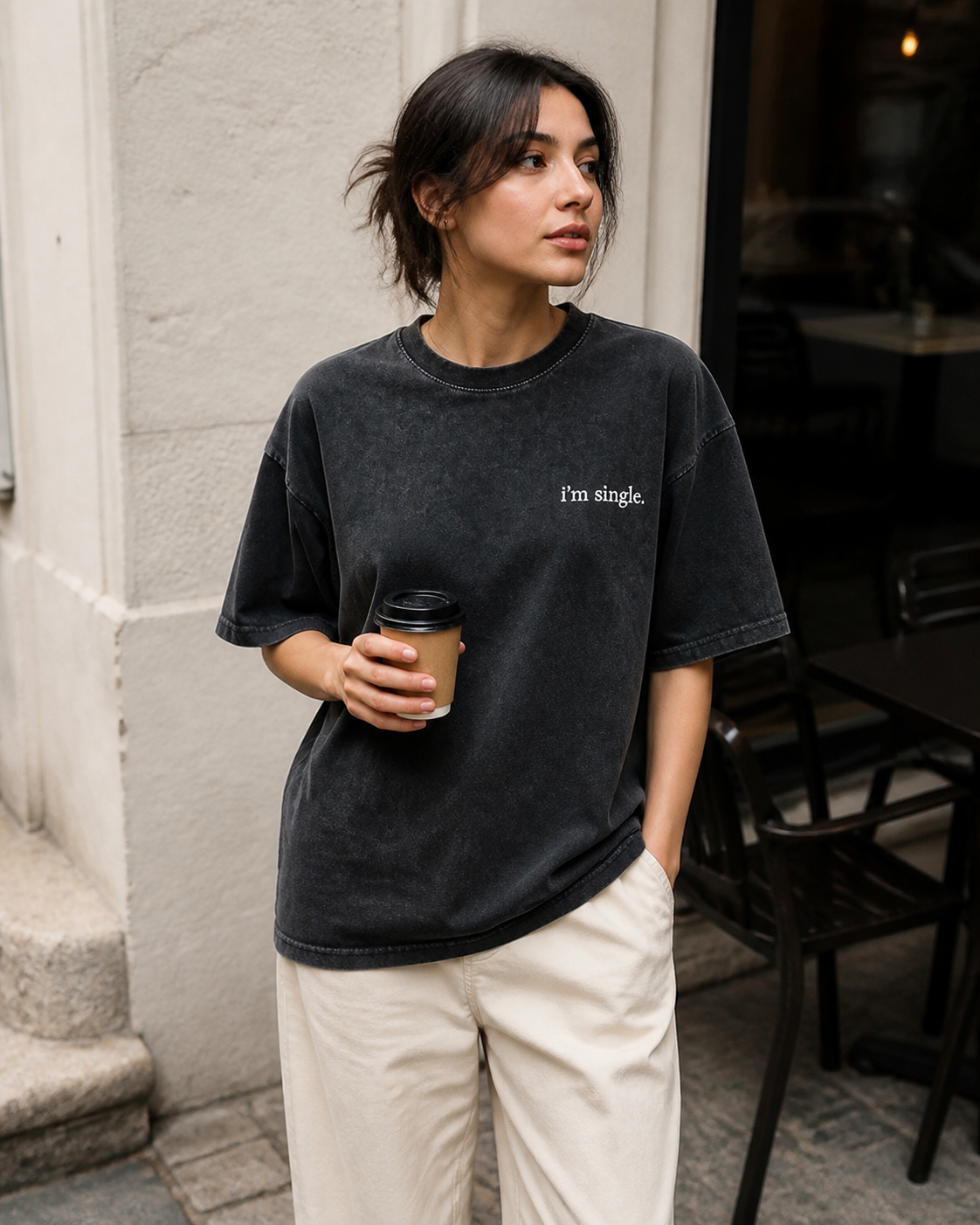 A woman wearing a washed black graphic t-shirt with the text 'I'm single.' on the chest, paired with cream-colored pants, holding a coffee cup in an outdoor cafe setting.