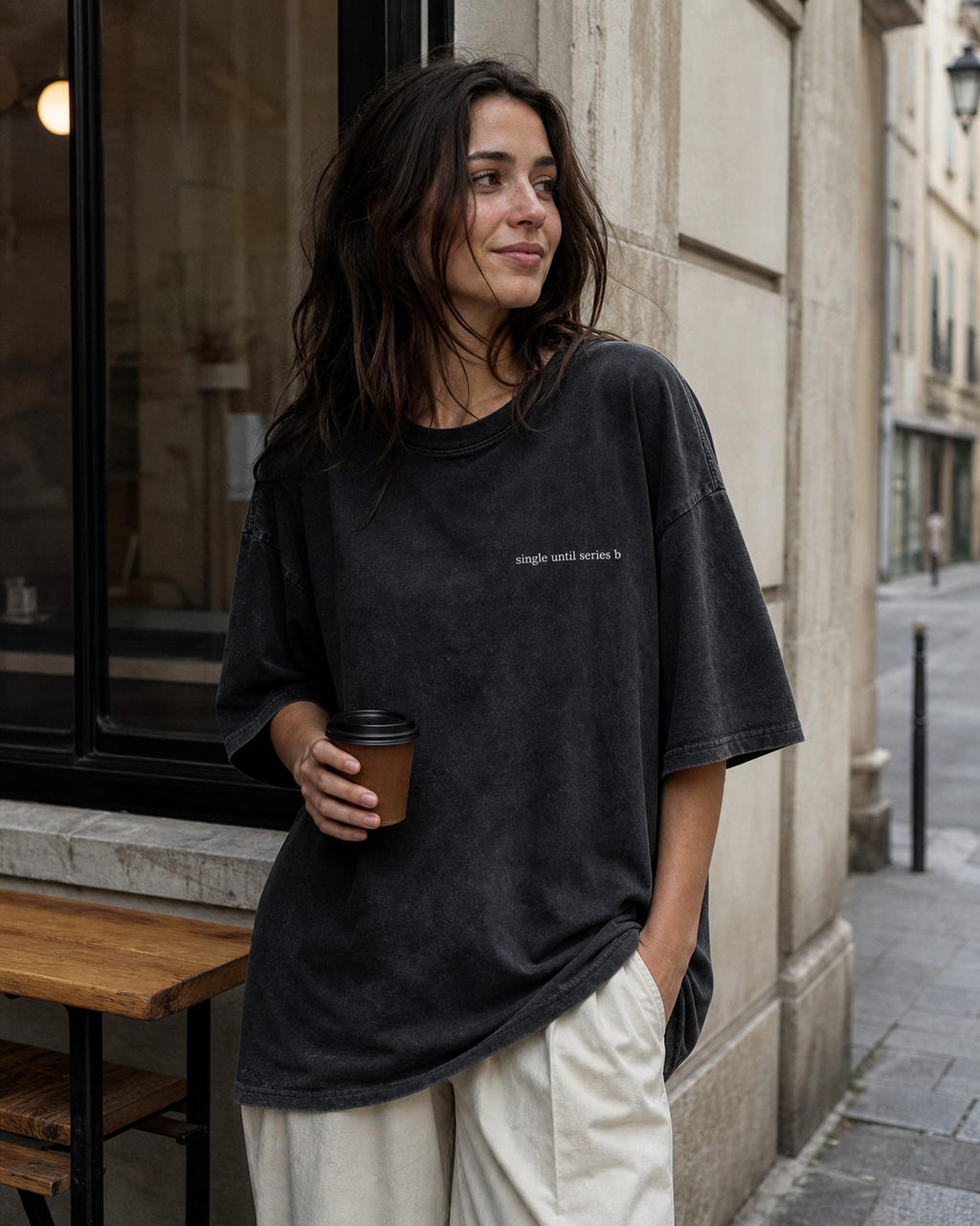 A woman stands on a city street wearing an oversized faded black t-shirt with small white text on the chest, paired with cream-colored pants, holding a brown paper coffee cup.