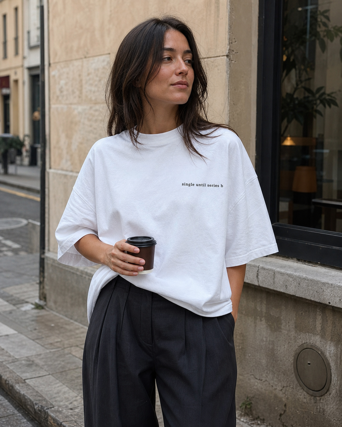 A woman on a city street wearing an oversized white t-shirt with small black text on the chest, paired with cream-coloured trousers, holding a paper coffee cup.