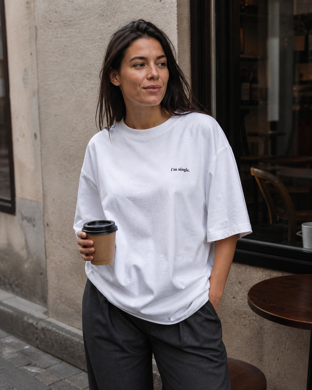 A woman wearing a white graphic t-shirt with 'i'm single.' printed in black on the chest, paired with cream-coloured trousers, holding a coffee cup in an outdoor cafe setting.