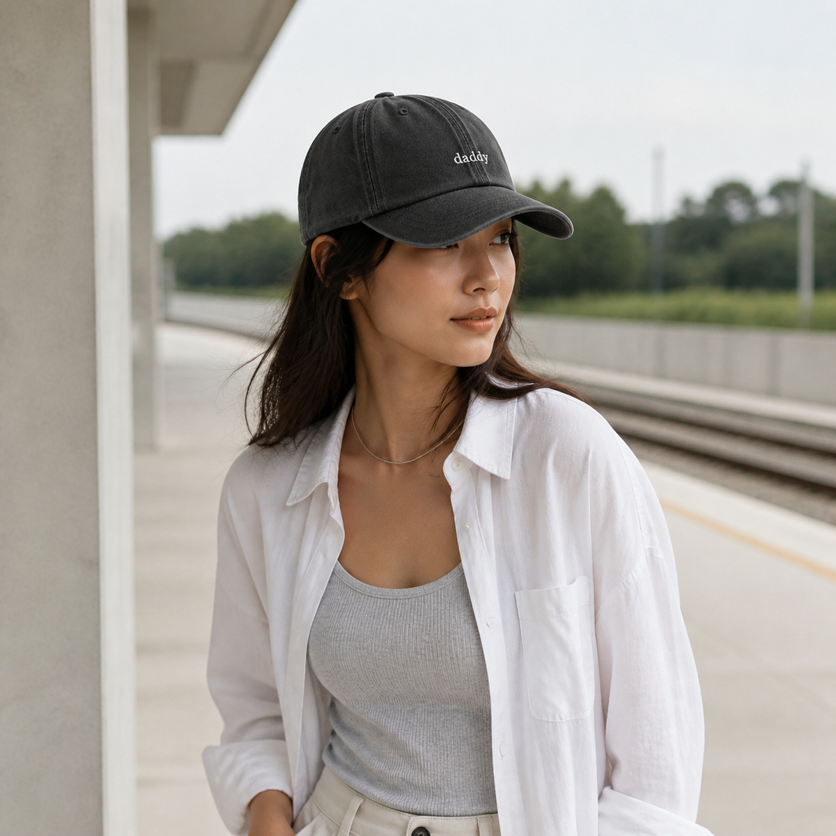 A woman wearing a dark gray baseball cap with the brand name 'daddy' embroidered in white on the front, styled with a white button-up shirt and a light gray ribbed tank top, standing on a train platform.
