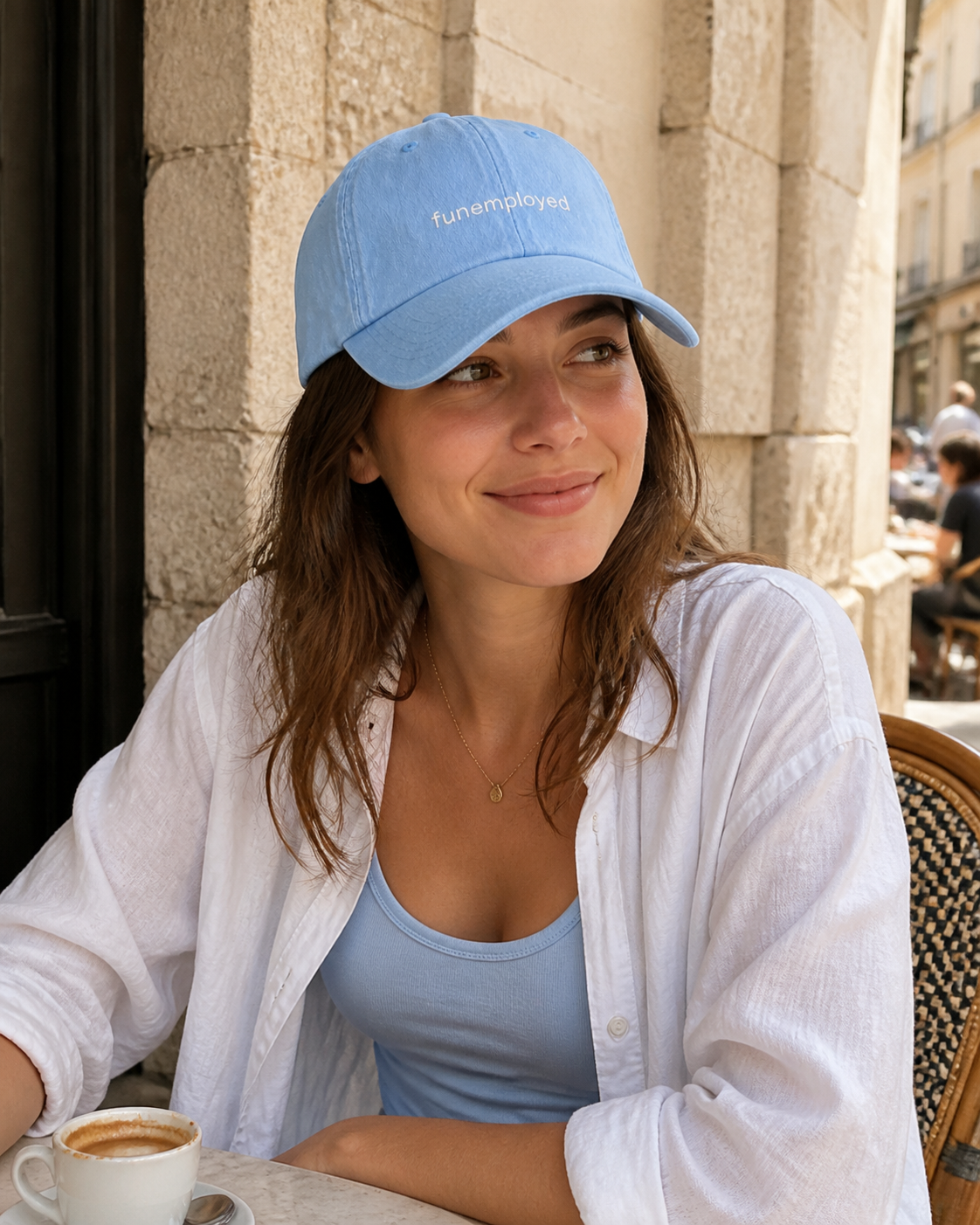 A woman sitting at a cafe table wearing a light blue baseball cap with the text 'fun.employees.
