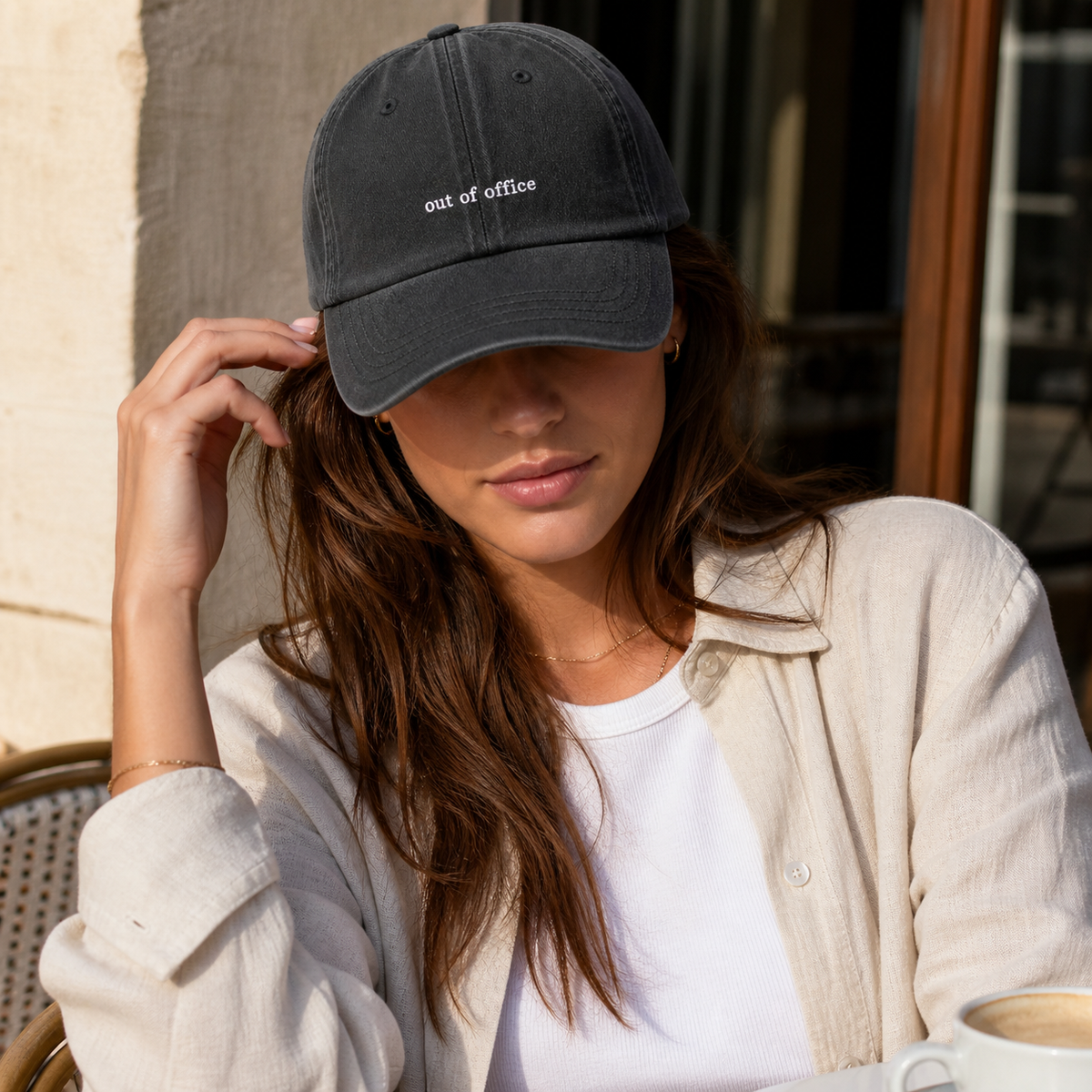 A woman wearing a dark charcoal gray baseball cap with the words 'out of office' embroidered in white on the front, styled with a beige button-up shirt and white t-shirt.