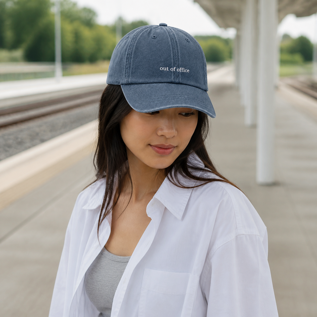 A woman wearing a washed blue baseball cap with 'out of office' embroidered in white, standing on a train station platform.