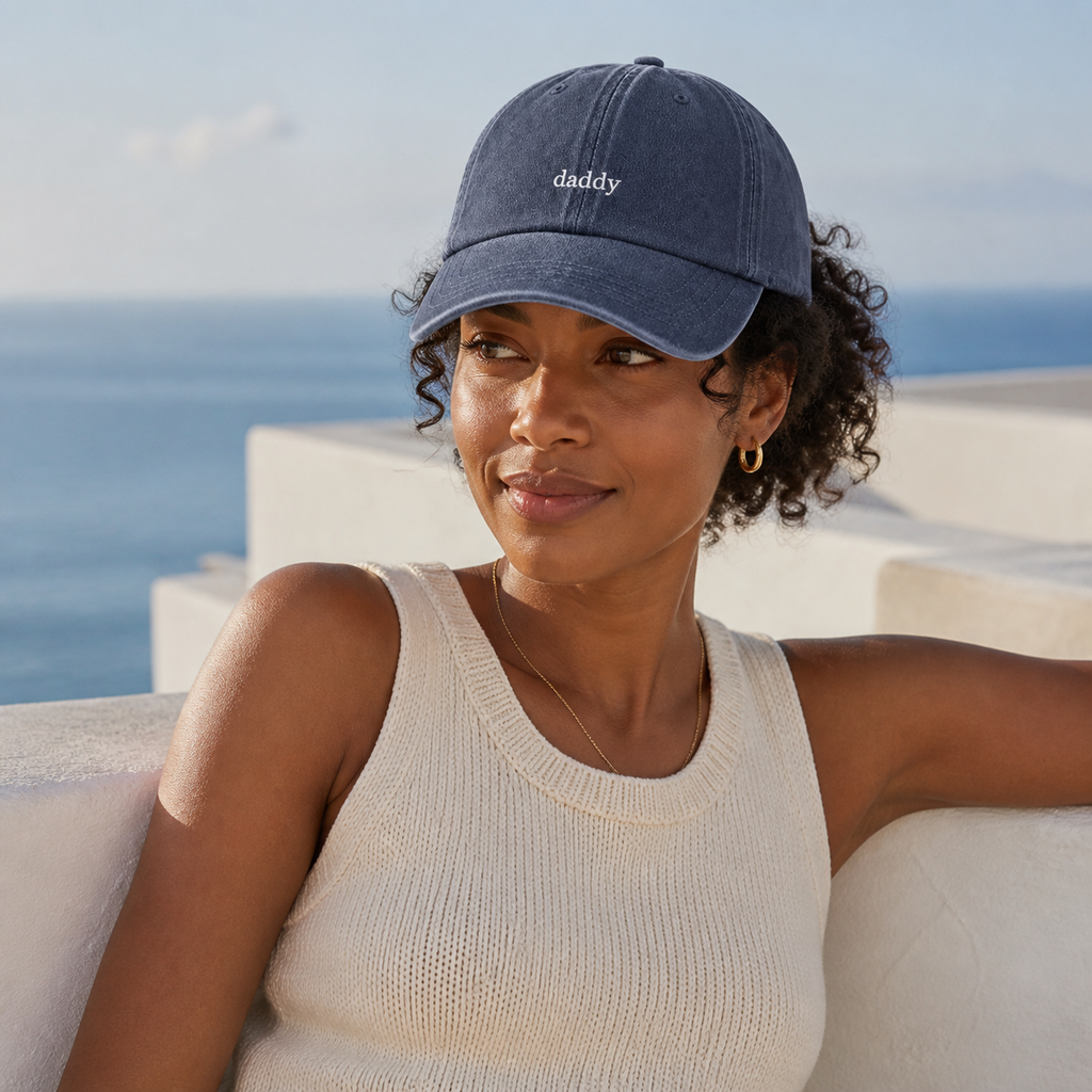A woman wearing a navy blue baseball cap with 'daddy' embroidered in white on the front, posing outdoors with the sea and white buildings in the background.