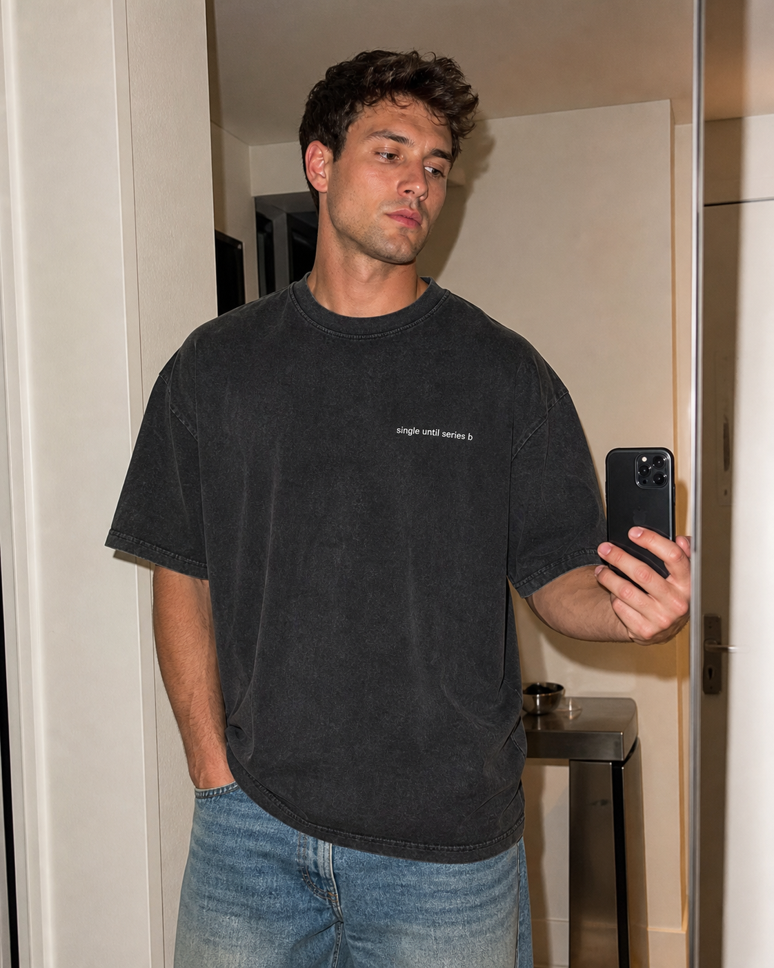 A man taking a mirror selfie wearing a washed black graphic t-shirt with 'simple love everything' printed on the chest, paired with blue jeans.