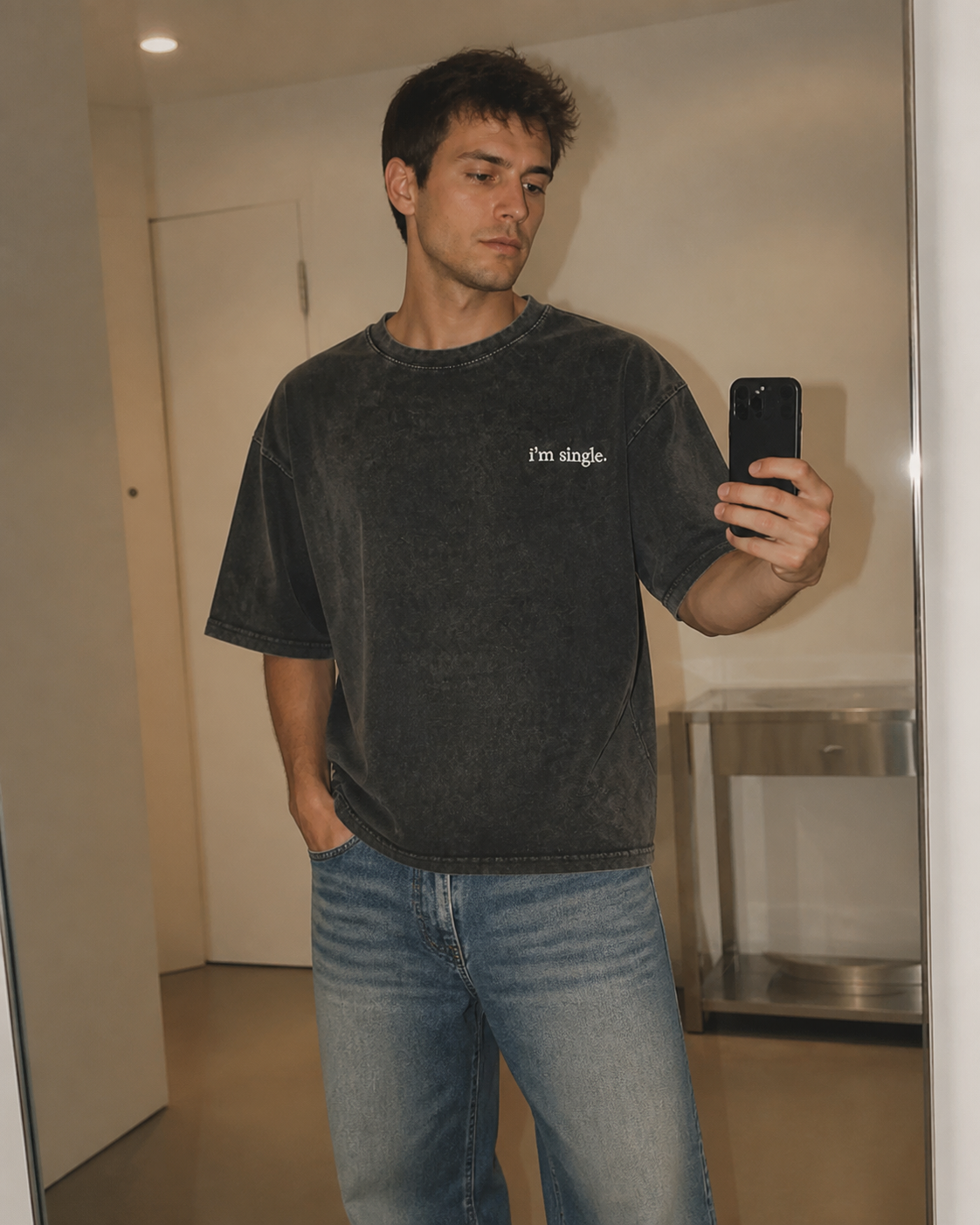 A man taking a mirror selfie wearing a washed black t-shirt with the text 'I'm single.' on the chest and blue jeans.