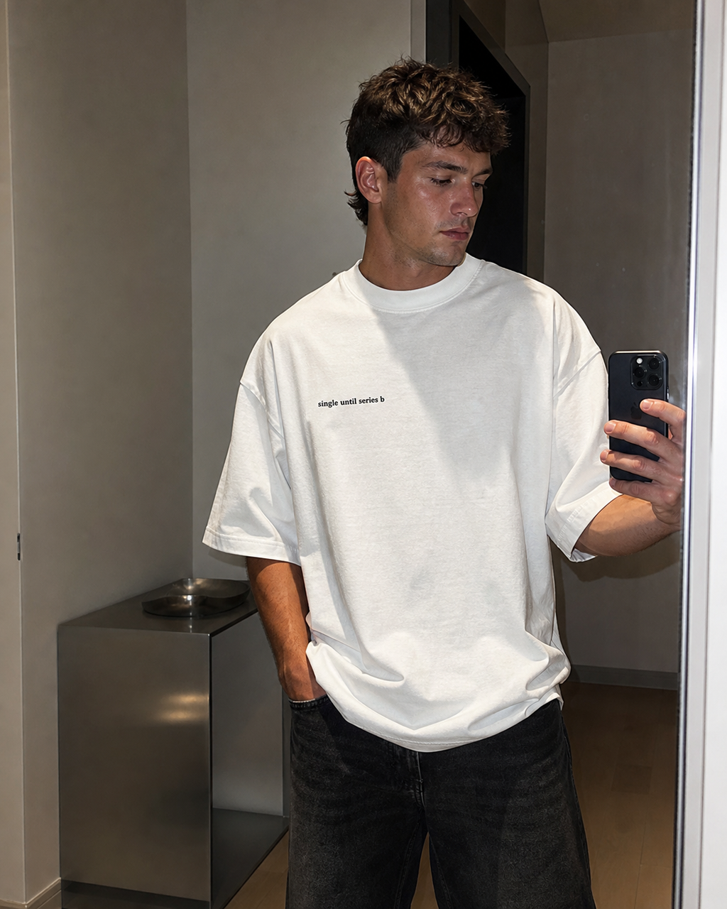 A man taking a mirror selfie wearing a white graphic t-shirt with small black text on the chest, paired with blue jeans.