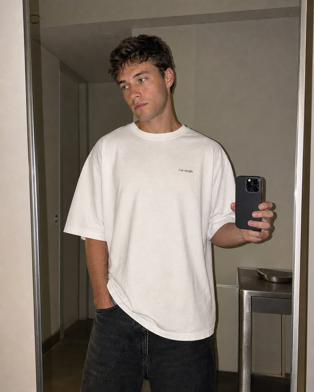 A man taking a mirror selfie wearing a white t-shirt with 'i'm single.' printed in black on the chest, paired with blue jeans.