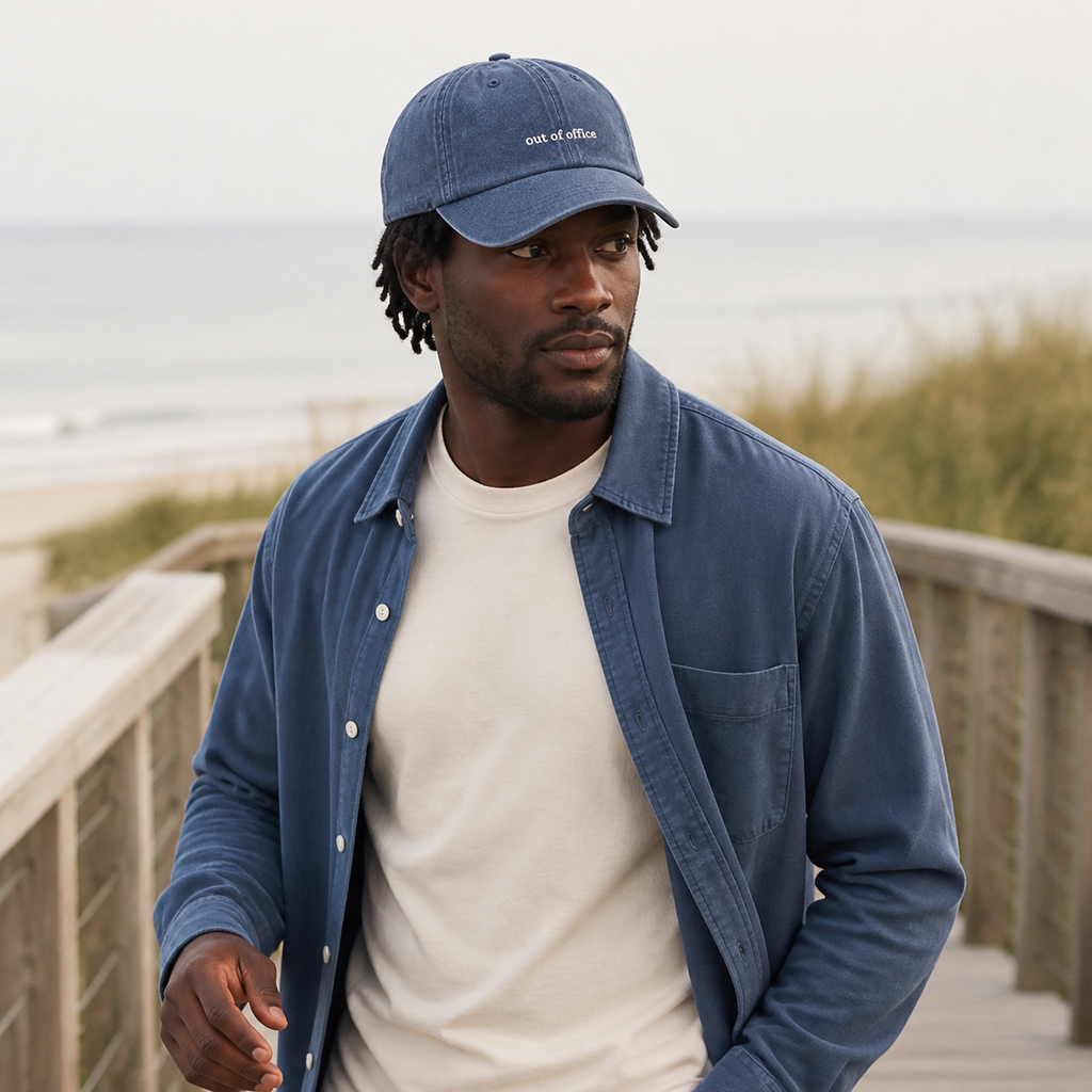 A man wearing a blue denim baseball cap with 'one of these' embroidered on the front, a blue button-up shirt, and a cream t-shirt, standing on a wooden boardwalk with a beach and ocean blurred in the background.