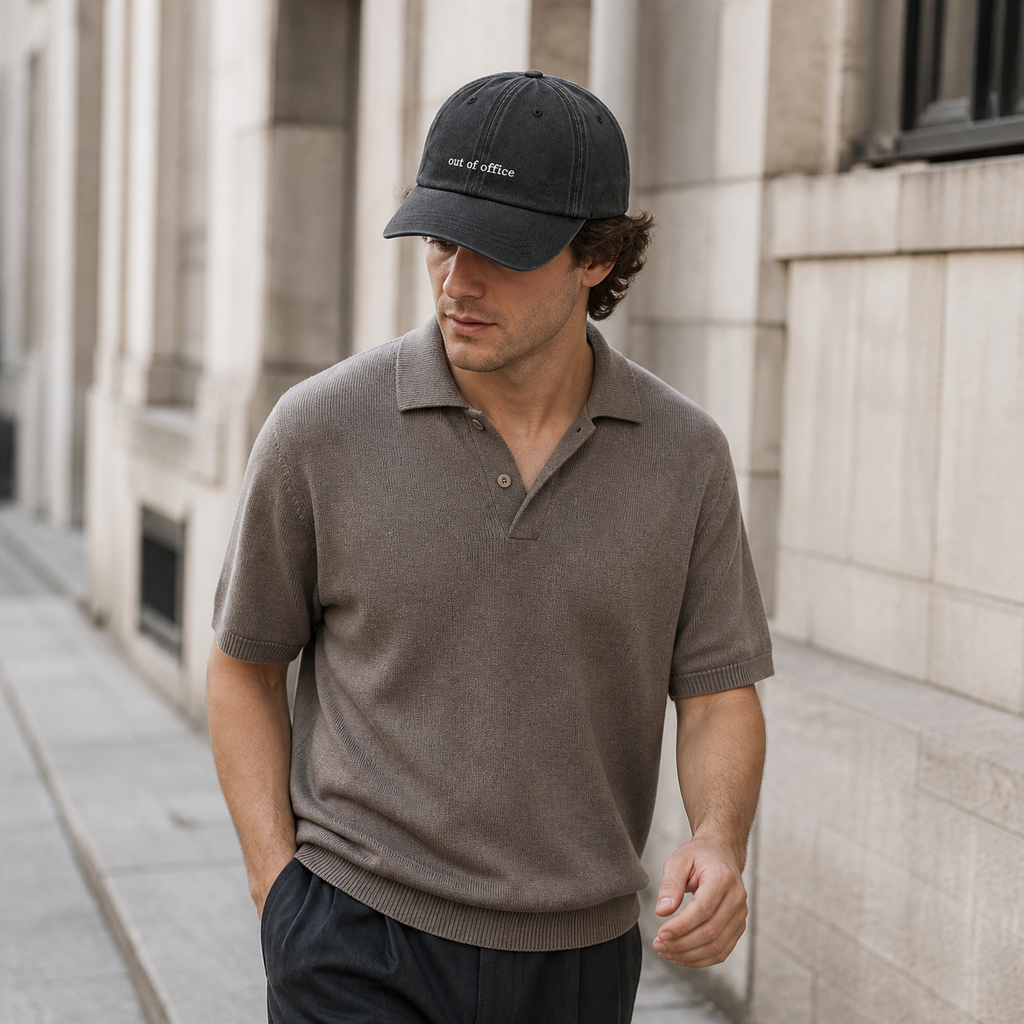 A man wearing a dark gray baseball cap with 'out of office' embroidered on the front and a taupe short-sleeve knit polo shirt, walking outdoors against a light stone building.