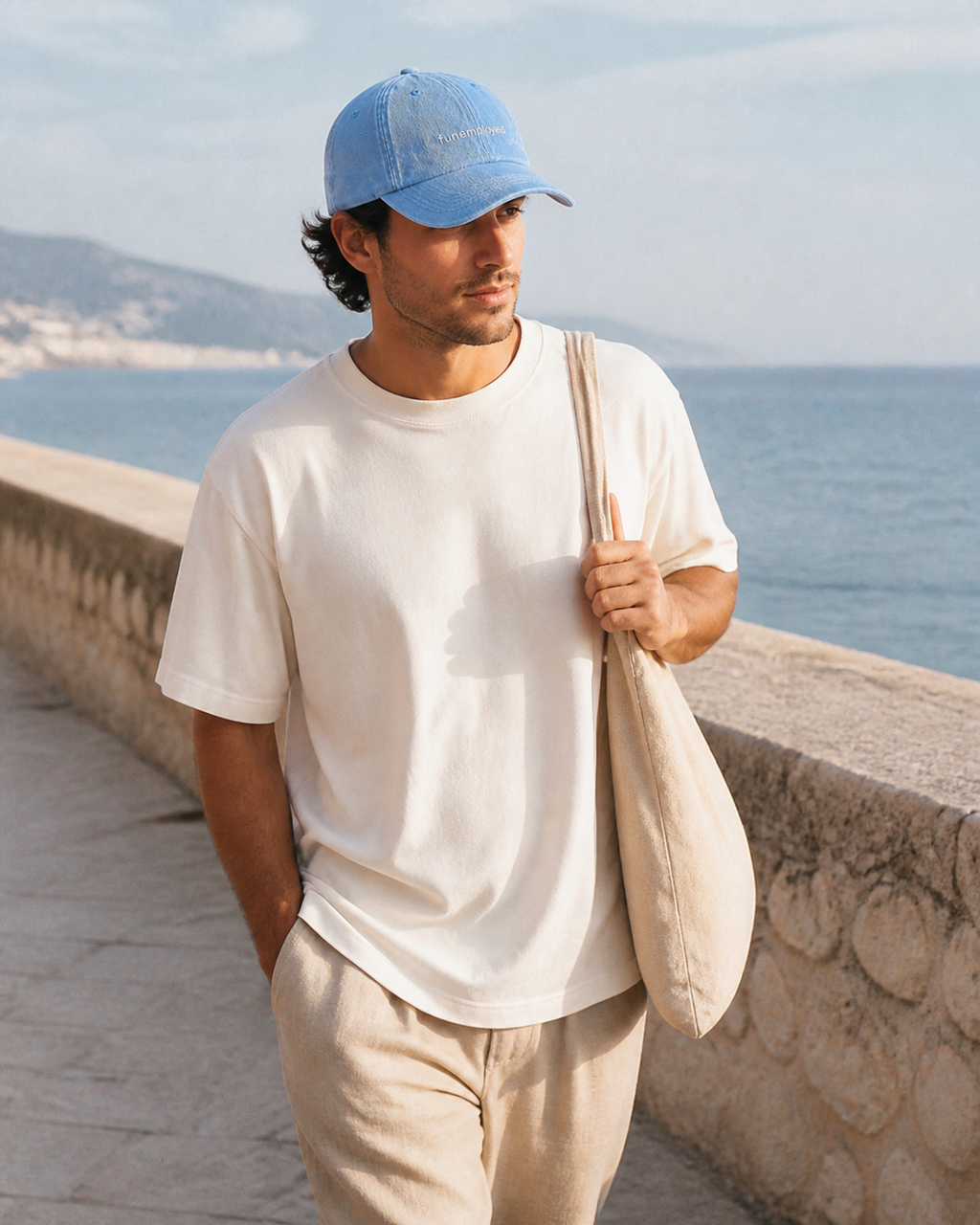 A man wearing a light blue baseball cap, a cream-colored t-shirt, and beige pants walks along a stone path by the sea, carrying a beige canvas shoulder bag.