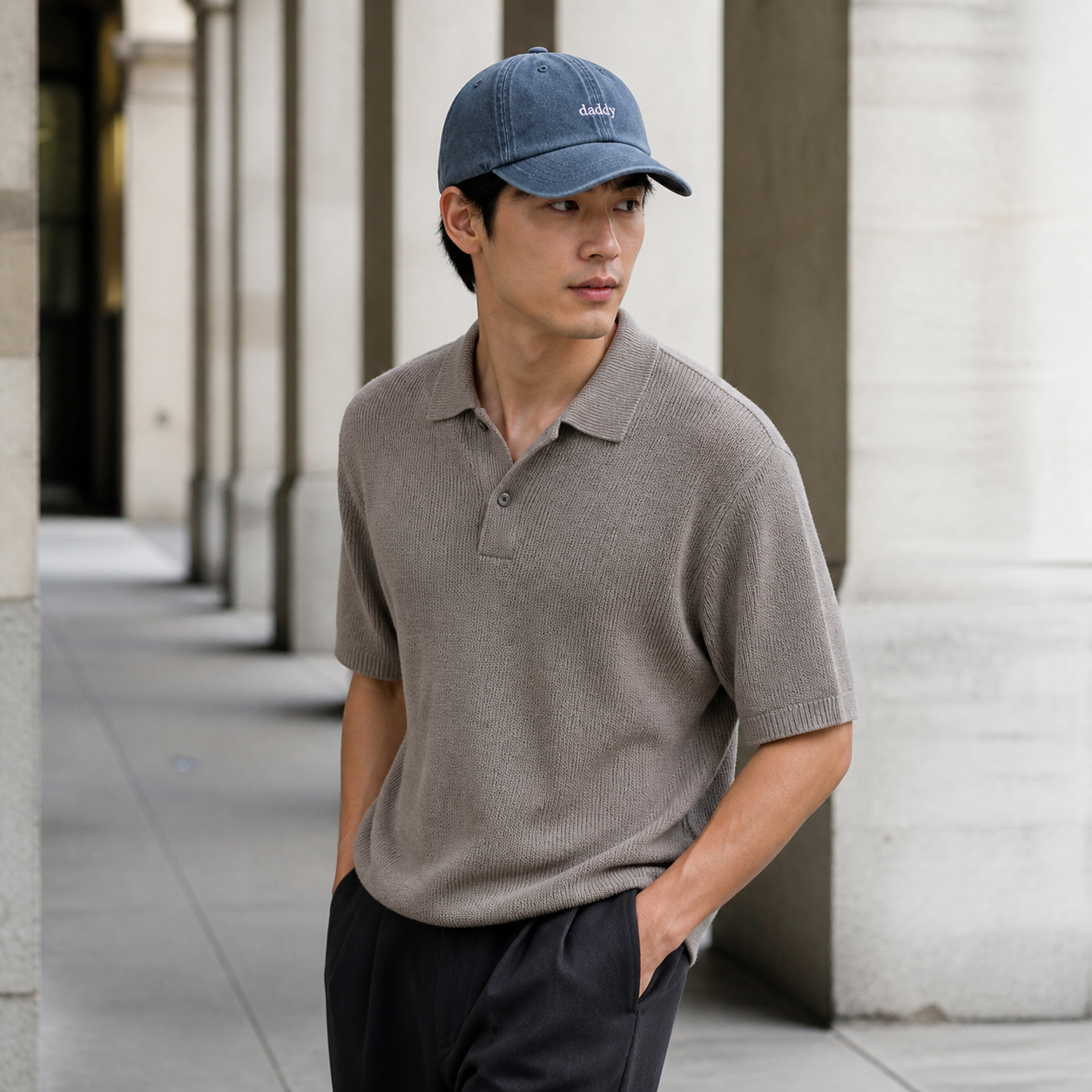 A man wearing a slate blue baseball cap with 'd&b&' embroidered on the front, a taupe short-sleeve knit polo shirt, and dark trousers, standing in a modern outdoor walkway with columns.